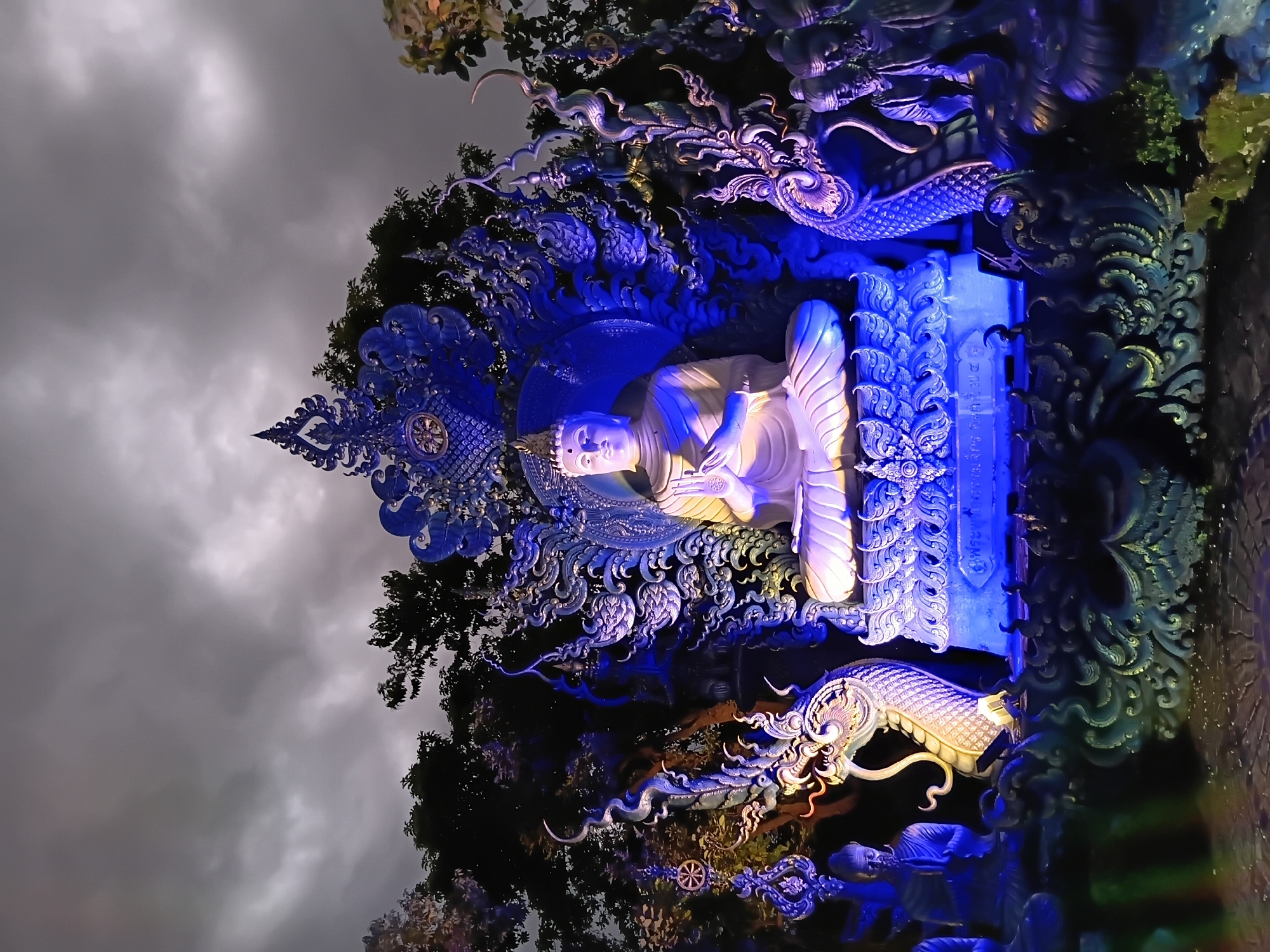 This image depicts the exterior of the Blue Temple (Wat Rong Suea Ten) in Chiang Rai City Municipality, Thailand, at night. The central subject is a large, white Buddha statue seated in a meditative pose, illuminated with blue and white lights. It is surrounded by intricate, ornate blue sculptures resembling mythical serpents or dragons, with detailed scales and expressive faces. Behind the Buddha statue is a large, elaborate blue headdress or backdrop, also adorned with decorative elements. To the left and right of the central Buddha, smaller statues of robed figures in prayer are visible. The overall scene is bathed in dramatic blue and purple lighting, casting shadows and highlighting the detailed craftsmanship of the sculptures. Trees with dark foliage are visible in the background against a cloudy, dark sky. The ground appears to be paved.