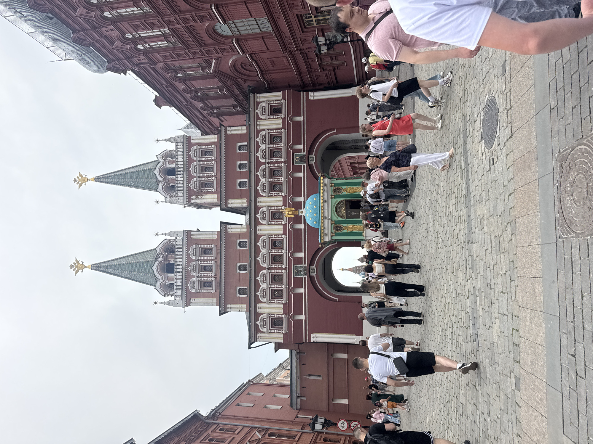 The image depicts a scene at Red Square in Moscow, Russia. A large, ornate, red brick building with decorative white trim and multiple windows dominates the center background. Two tall, green-roofed towers with golden double-headed eagles on top rise from this building. In front of the central archway of the building, there is a smaller, colorful structure with a blue and gold dome. Numerous people, dressed in casual summer clothing, are walking on a cobblestone plaza in front of the building. The sky is overcast.