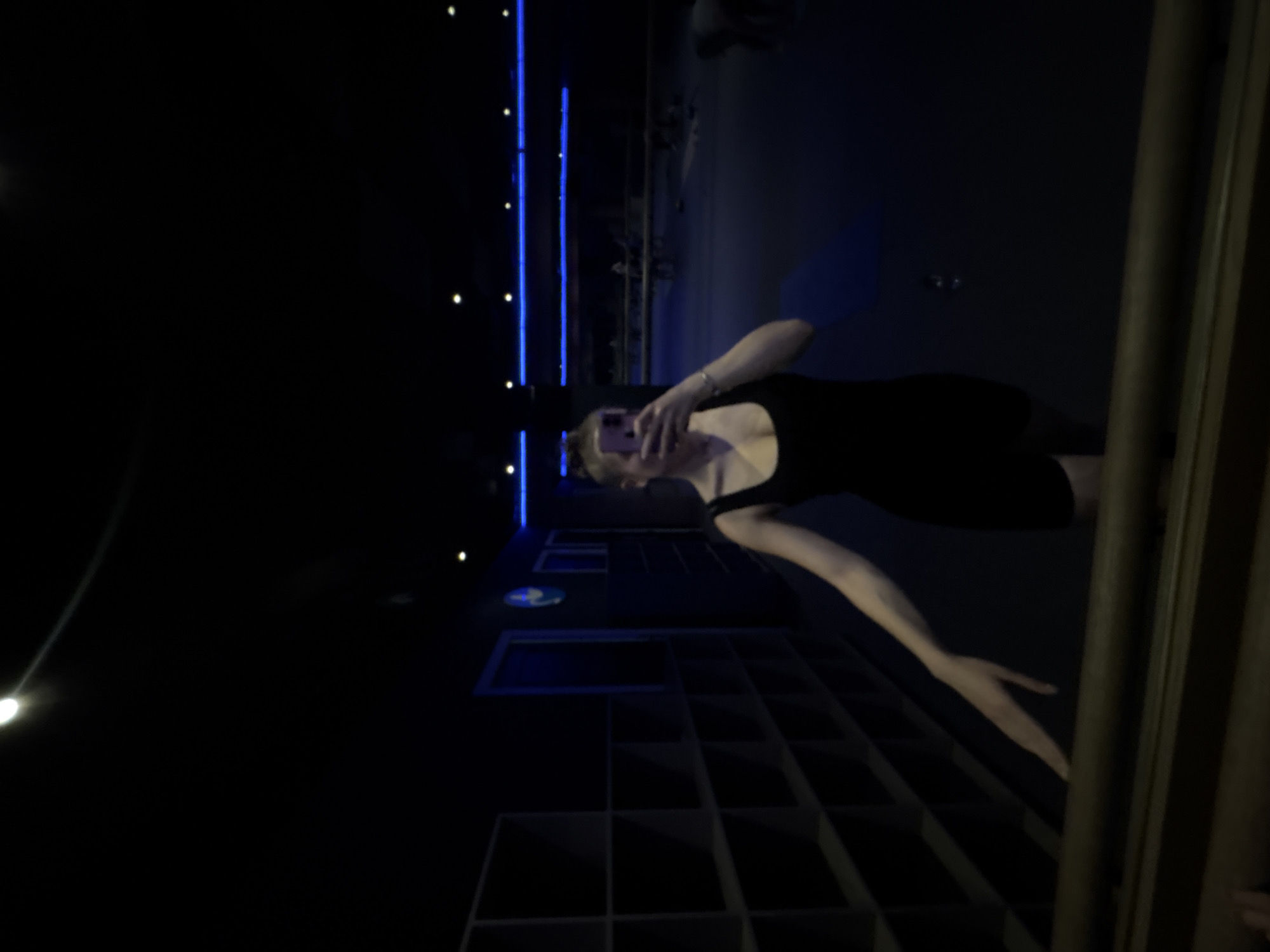 A person wearing a black outfit stands on a staircase in what appears to be an indoor setting. The person is holding a mobile phone up, presumably taking a selfie. The staircase has a grid-like design, and there are railings visible on the right side. The location is in Mexico City, Mexico. The environment is dimly lit, with blue light accents.