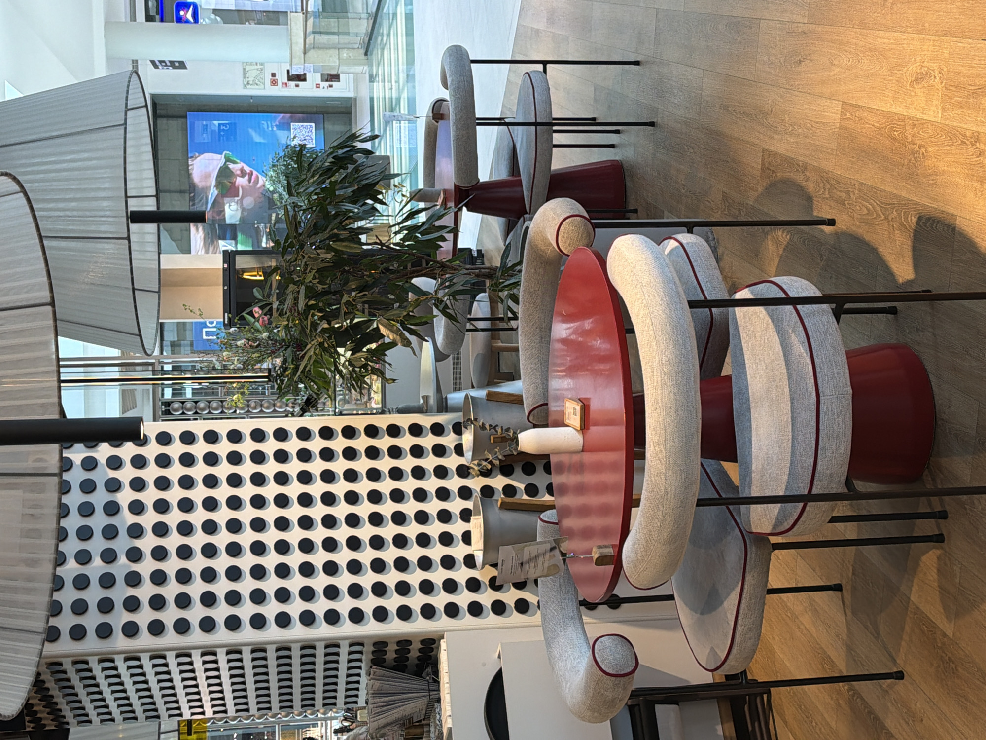 This media file captures a modern, unoccupied seating area, likely a cafe or lounge, within a commercial building in Khimki, Russia. The image appears to be taken in portrait orientation and rotated 90 degrees clockwise.
The scene is dominated by several sets of stylish furniture. Each set includes a circular, glossy red table and light grey upholstered chairs with distinctive curved backs and arms, accented with red piping. The chairs rest on slender black metal frames. On the tables, there are small white cups or containers, and some have small, unreadable labels attached.
To the left, large, translucent white, ribbed cylindrical lamps or light shades with black support poles are visible. A vibrant artificial plant with green leaves and a few small flowers stands between the seating arrangements. Behind the plant and furniture, a white wall features a striking pattern of regularly spaced black circular disks.
In the background, a large digital display screen shows an image of a person wearing sunglasses, accompanied by some white text and a QR code, none of which are clearly legible. Beyond the screen, more of the building's interior is visible, including glass railings and other structures, suggesting a multi-level environment like a shopping mall or atrium.
The floor is made of light-colored wooden planks, showing natural wood grain patterns and reflections from overhead lighting. The overall aesthetic is clean and contemporary. No people are present, so there is no specific activity taking place. The indoor setting means the time of day and weather are not discernible.