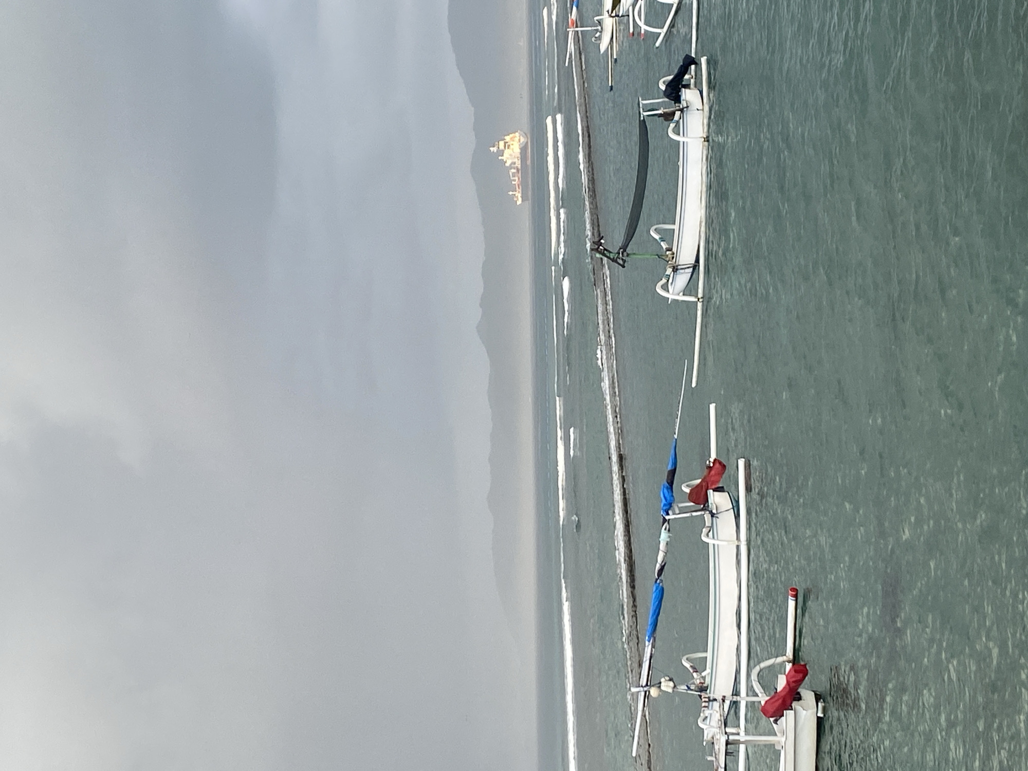 A wide shot shows a calm, gray ocean with a coastline in the distance. Several traditional outrigger canoes are floating in the foreground and mid-ground. The sky is overcast, with a hint of sunlight illuminating a large cargo ship on the horizon. The water is a deep blue-green, with gentle waves lapping against the shore. The scene appears to be set in Karangasem, Indonesia, a coastal town known for its tranquil beaches and traditional fishing villages.