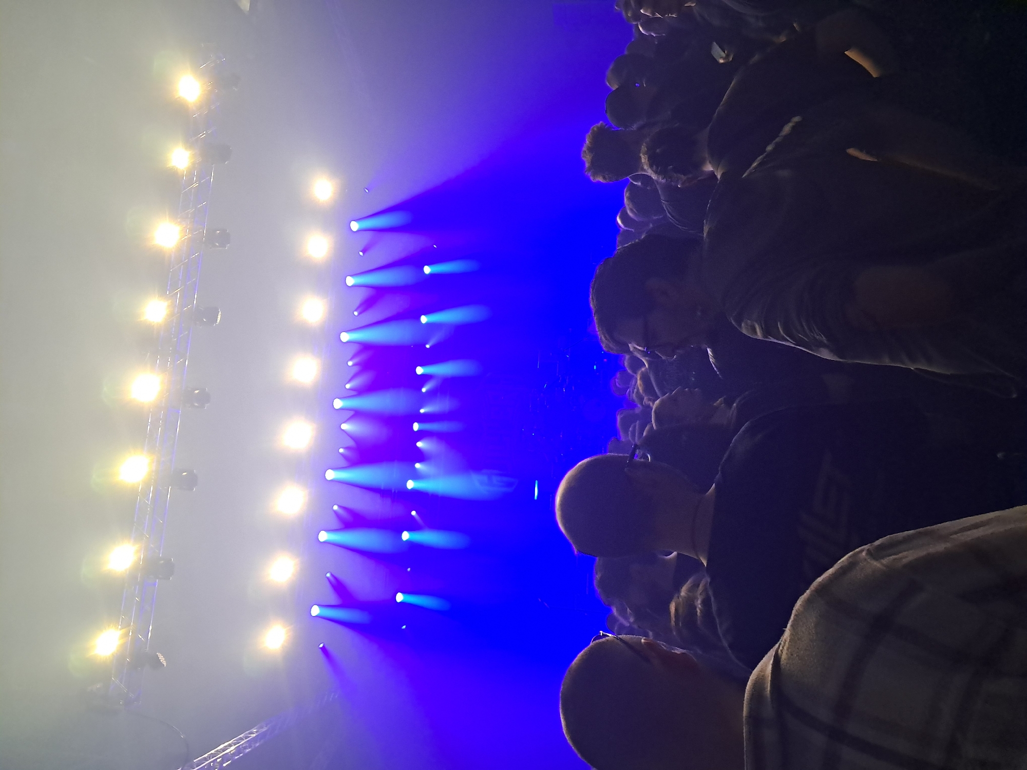 The image depicts a crowd of people attending a live music concert. The location is likely an indoor venue in Sofia, Bulgaria based on the architecture and event aesthetics common in the city. Several rows of lights emit bright white and blue beams across the space, creating a dynamic visual effect. The crowd is dense, with people standing closely together and facing towards the stage, suggesting they are watching a performance. The lighting creates a smoky atmosphere with a distinct stage presence.