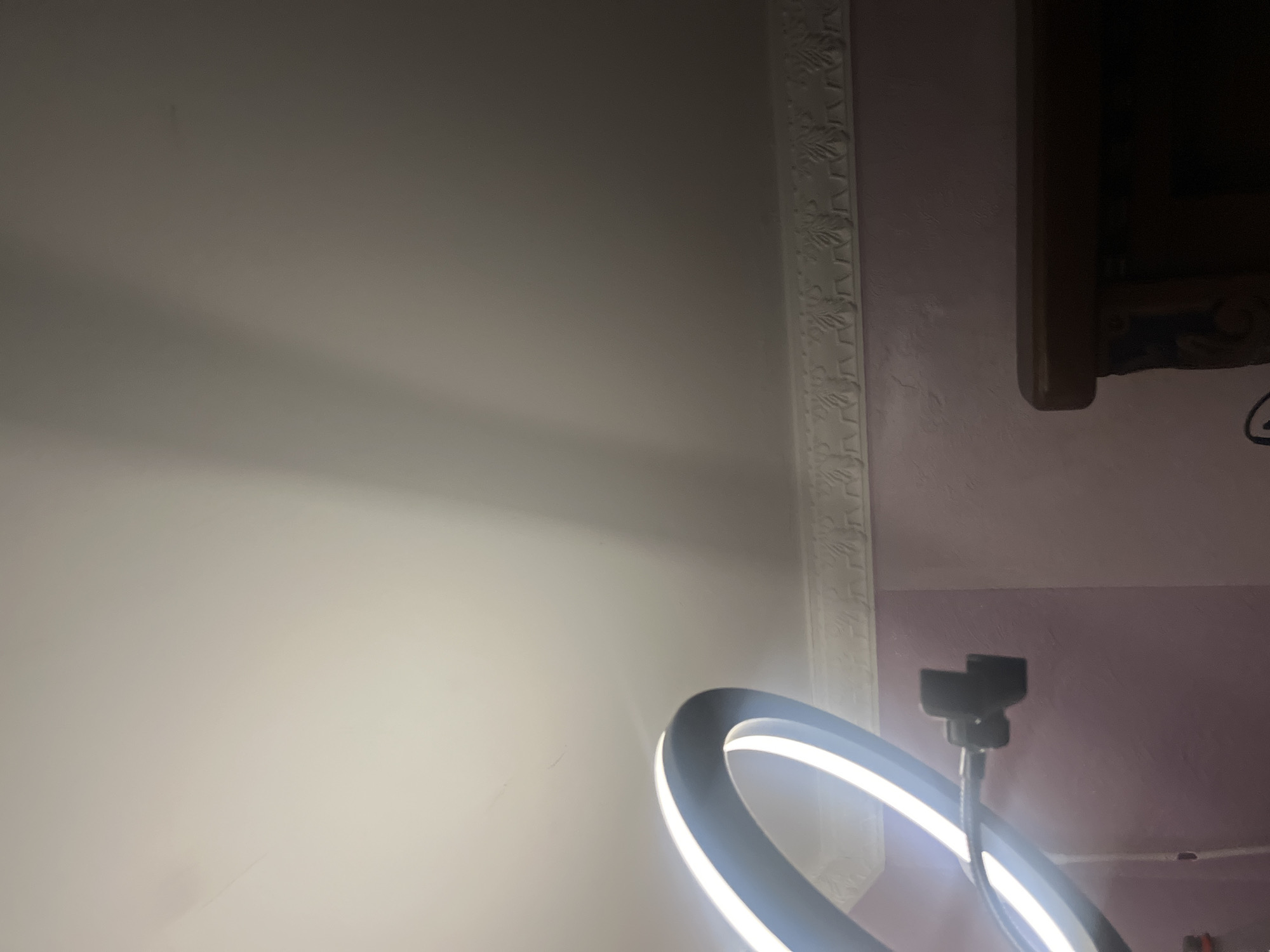 The image shows a dimly lit room with a pink textured wall and a white ceiling. A ring light is partially visible in the lower left corner, casting bright light onto the ceiling. A decorative molding runs along the top of the wall, below the ceiling. In the lower right corner, the top of a dark wooden piece of furniture, possibly a cabinet or shelf, is visible. The overall scene suggests an indoor setting, likely a room within a residence. There are no people visible in the image.