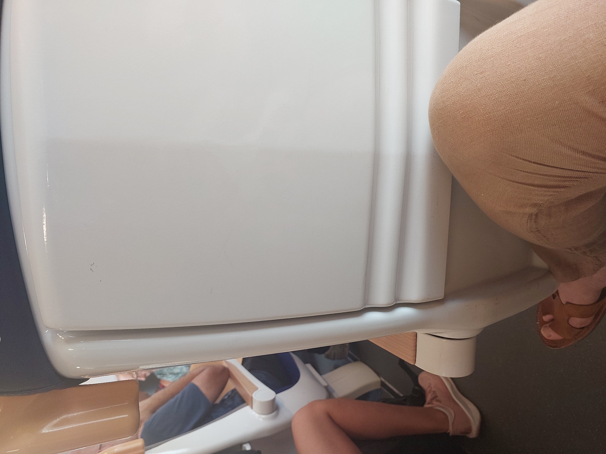 The image shows a close-up view of people seated on a form of public transportation, likely a train or bus, due to the rows of seats. The perspective is from a seated passenger looking forward and slightly to the side. In the foreground, the legs and feet of two individuals are visible. One person is wearing tan sandals and has their legs crossed, with their right foot resting on the floor. The other person, slightly further back and to the left, is wearing pink sneakers and has their legs bent, with their right leg visible from the knee down.
Behind these individuals, glimpses of other passengers and the seating arrangement can be seen. The seats appear to be modern, with white plastic backs and wooden armrests. The interior lighting is somewhat dim, with the white plastic of the seats reflecting light. The environment is clean and functional, typical of public transit. The overall scene suggests a journey in progress, with people in transit. The location is not specified beyond "Unknown, Unknown," and there are no city-specific landmarks or identifiers. The image captures a mundane moment of everyday travel.