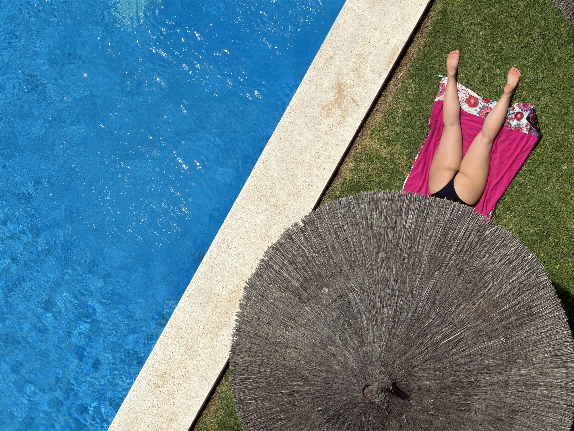 An aerial shot captures a scene by a swimming pool on a bright, sunny day. The vibrant blue water of the pool takes up the left half of the frame, its surface rippling and reflecting the sunlight. A beige-colored tiled border separates the pool from the lush green grass on the right. On the grass, a person is lying on their back on a pink towel with a floral pattern. They are wearing black bikini bottoms and their legs are propped up in the air. Partially obscuring the foreground is a large, straw-covered sun umbrella, its textured surface creating a circular shape. The scene evokes a sense of relaxation and leisure, suggesting a vacation or a warm, leisurely day outdoors. The lighting indicates it is daytime. There is no visible text in the image.