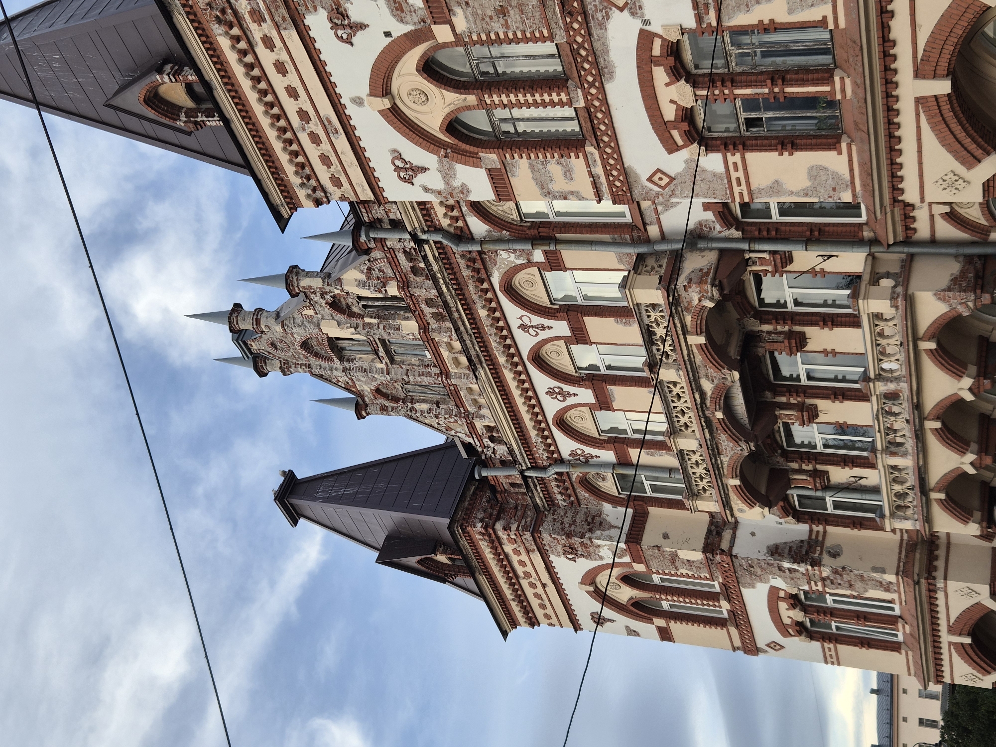 The image shows the facade of an old building in Vyborg, Russia. The building is made of brick and stone, with a distinctive architectural style featuring arched windows, decorative cornices, and pointed towers. The facade shows signs of wear and tear, with chipped paint and exposed brickwork. The sky is cloudy with a few streaks of blue. There are power lines visible in the foreground. The building appears to be multi-story, with several windows and balconies. The architectural style suggests it may be a historical building, possibly from the late 19th or early 20th century, common in the Vyborg region. The overall impression is of an aged but still impressive structure against a backdrop of a cloudy sky.