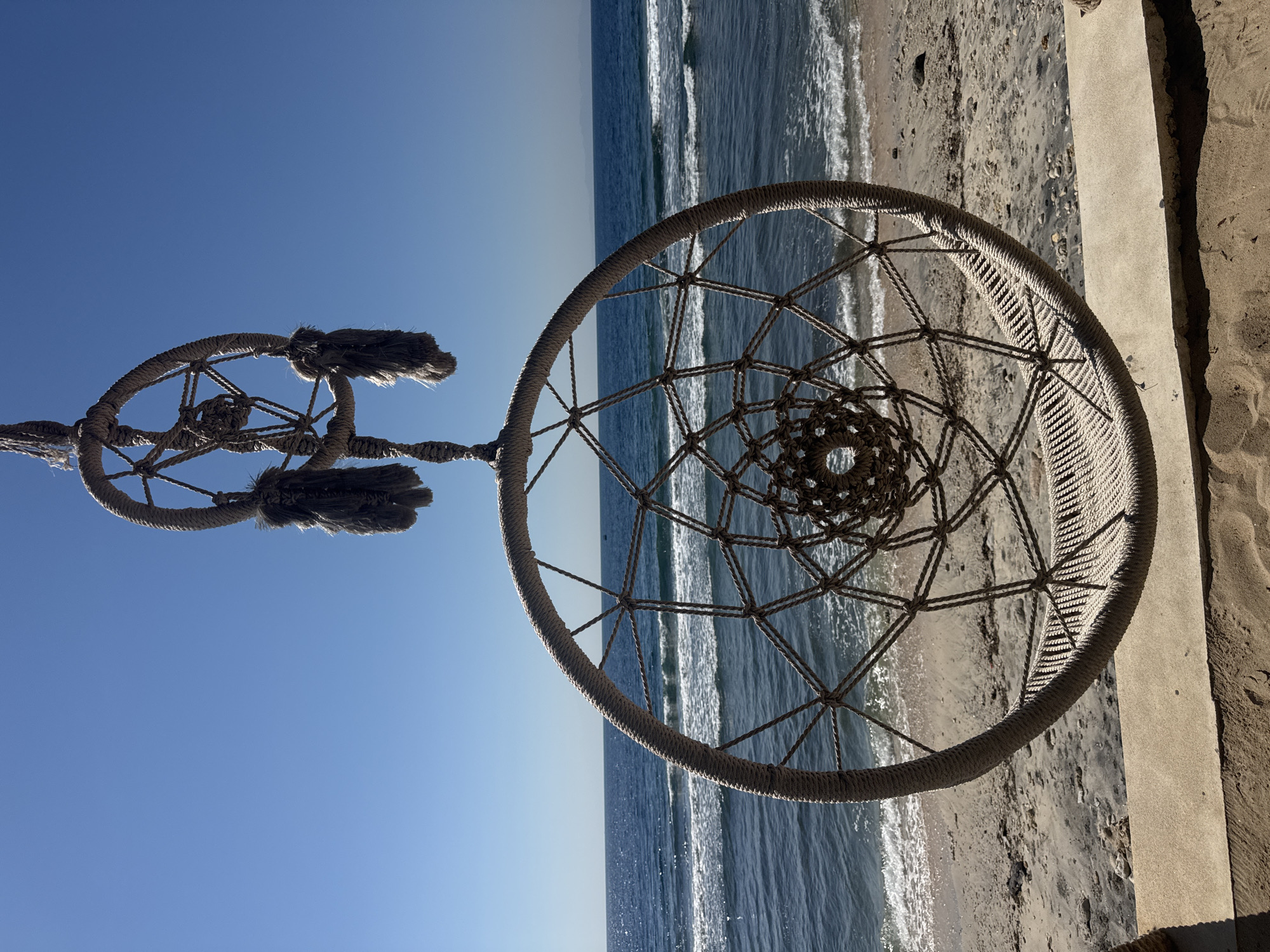The image captures a coastal scene in Nusa Penida, Indonesia, featuring two woven rope structures suspended in the foreground against a backdrop of the ocean and sky. The primary subject is a large, circular, open-weave macrame-style hanging structure, resembling a dreamcatcher or a swing seat, positioned centrally in the lower half of the frame. This structure is crafted from thick, natural-toned rope, intricately woven into a radial pattern with a denser, more supportive weave along its bottom curve. Directly above and partially overlapping this larger piece is a smaller, similar circular structure, also made of rope, from which several dark, shaggy tassels or fringes hang. Both structures are suspended from above by ropes extending out of the frame.
In the midground, the clear, deep blue waters of the ocean extend horizontally, meeting the horizon under a vast, cloudless light blue sky. Small, white-capped waves break gently near the shore, indicating calm water movement. A narrow strip of sandy beach, light tan with scattered darker patches and small rocks, lies between the water's edge and the foreground elements.
The immediate foreground features a light grey or tan concrete ledge running horizontally across the bottom of the frame. Beneath this ledge is more sand, displaying visible textures and shadows cast by the ledge itself and parts of the hanging structures, suggesting bright overhead sunlight. No individuals are present in the scene.