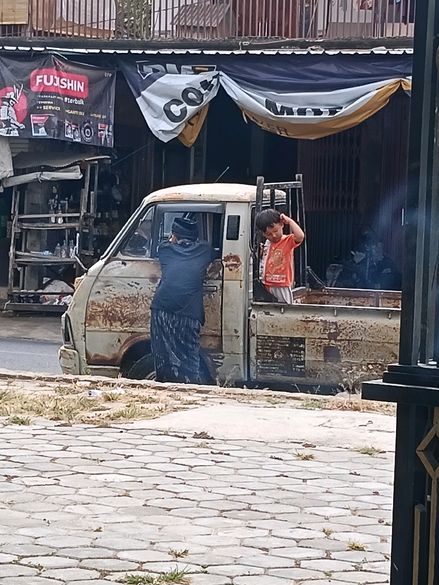In Bulangan Barat, Indonesia, a weathered, light-colored pickup truck with visible rust and damage is parked on a street. A man wearing a black cap and a dark long-sleeved shirt with a patterned sarong stands beside the driver's side door, leaning towards the truck. Inside the truck bed, a young boy in an orange t-shirt and light-colored shorts is looking towards the viewer, with his right hand raised to his head. The truck is positioned in front of a building with a corrugated metal roof, partially obscured by banners. One banner, predominantly black, displays the word "FUJISHIN" in large red letters, accompanied by graphics and text. Another banner, draped and rippling, shows large black letters that appear to read "COVER" on a yellow and white background. The foreground shows a cobblestone pathway with sparse dry grass and some scattered debris. The overall scene suggests a casual roadside stop or a commercial area.