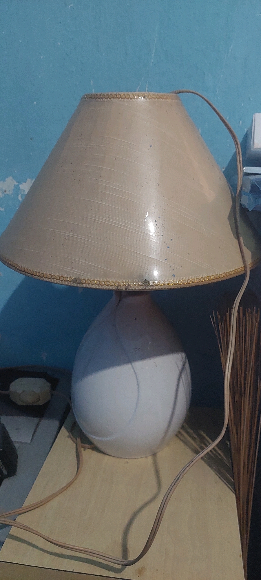 A lamp with a beige conical shade sits on a wooden surface against a light blue wall. The shade has a decorative gold trim around the bottom edge and appears to have some minor imperfections. The lamp base is a white, bulbous ceramic. A thin brown power cord snakes from the lamp base, crossing the wooden surface. To the right of the lamp, a bundle of dry, straw-like reeds stands upright. To the left of the lamp, a small, octagonal white object is visible, possibly a switch or a control, resting on a dark surface. The scene is indoors, likely in a domestic setting.