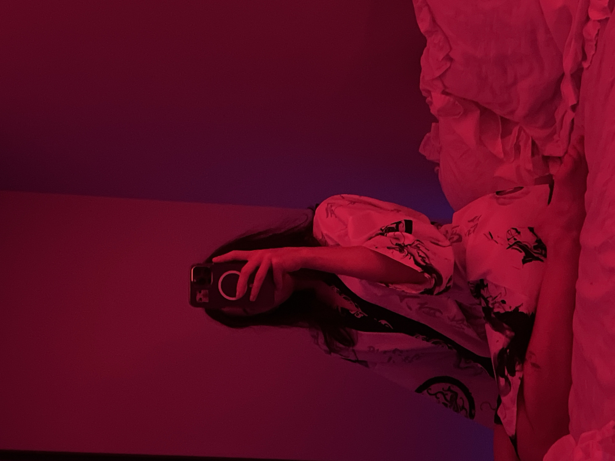 An individual, whose face is obscured by a smartphone held in their hand, is taking a mirror selfie. They are wearing a white robe with a black pattern and are seated on a bed with white, ruffled pillows. The scene is illuminated by red lighting, casting a strong hue over the environment. The background is a plain wall, also bathed in red light. The image was taken in Ternopil, Ukraine.