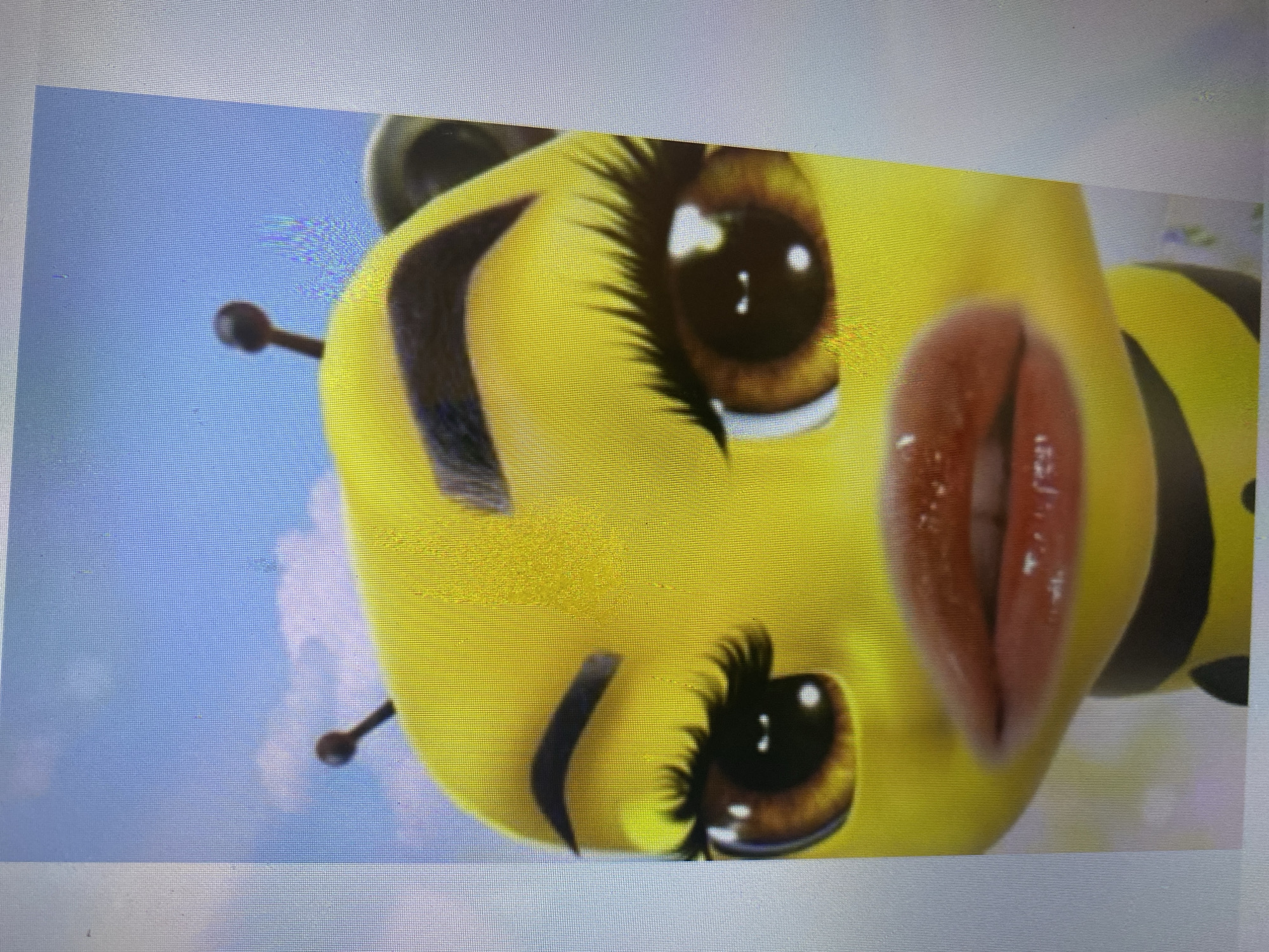 A close-up, head-and-shoulders shot features a stylized, anthropomorphic bee with exaggerated human-like features against a bright blue sky with wispy clouds. The bee has large, brown eyes with long, dark eyelashes and defined, arched eyebrows. Its lips are full and glossy red, and it has two small, dark antennae protruding from its head. The bee's body is yellow with black stripes. The image is a digital rendering, possibly from an animation or video game. The location in Ta', Indonesia is not discernible from the image.