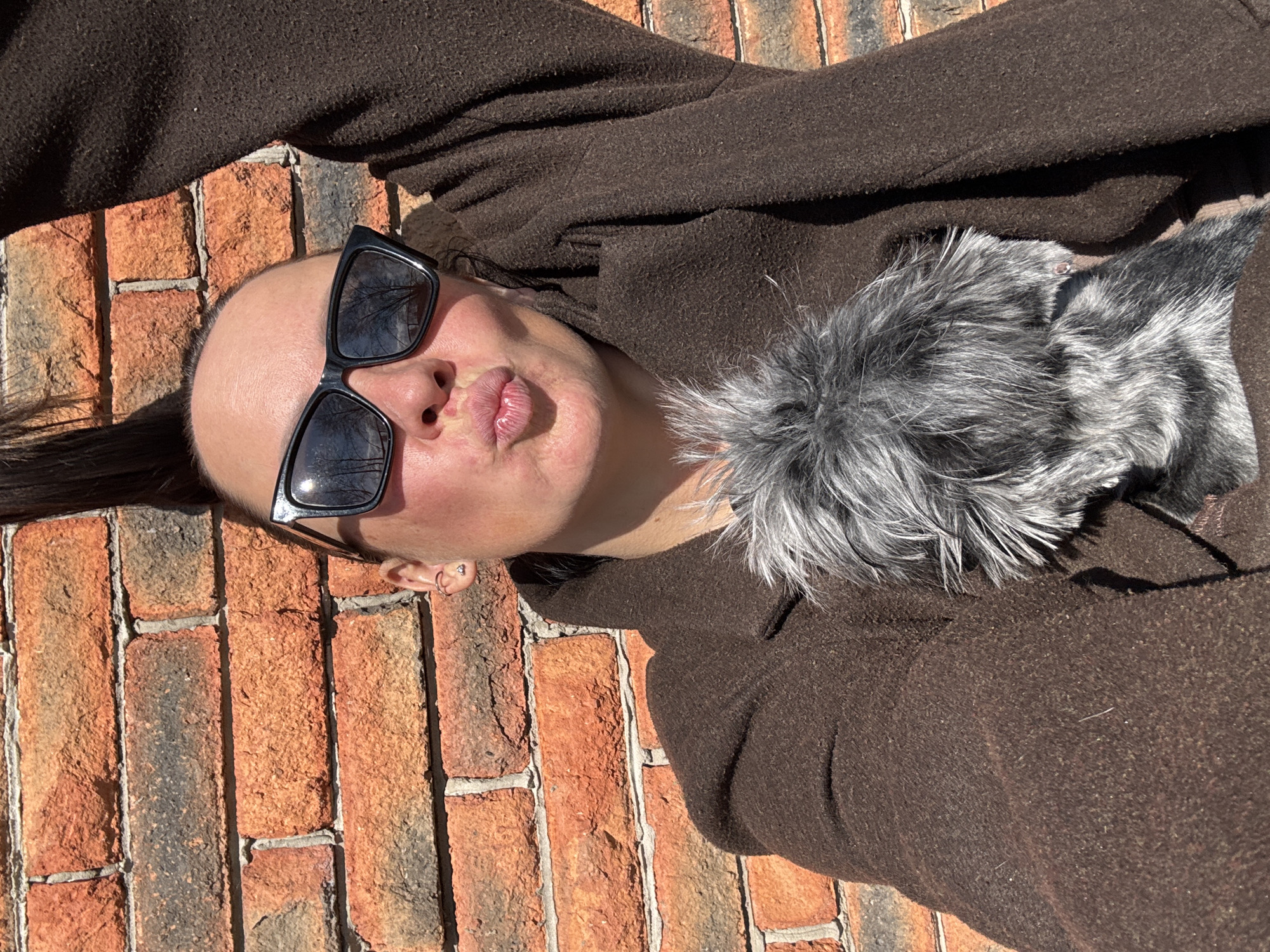 A person lies on a brick surface, possibly a wall or ground, wearing sunglasses and a brown coat with a fur collar. The person's face is turned towards the camera, with pursed lips. The location is unknown, and there's no visible evidence to suggest it's Chaikovsky, Russia.