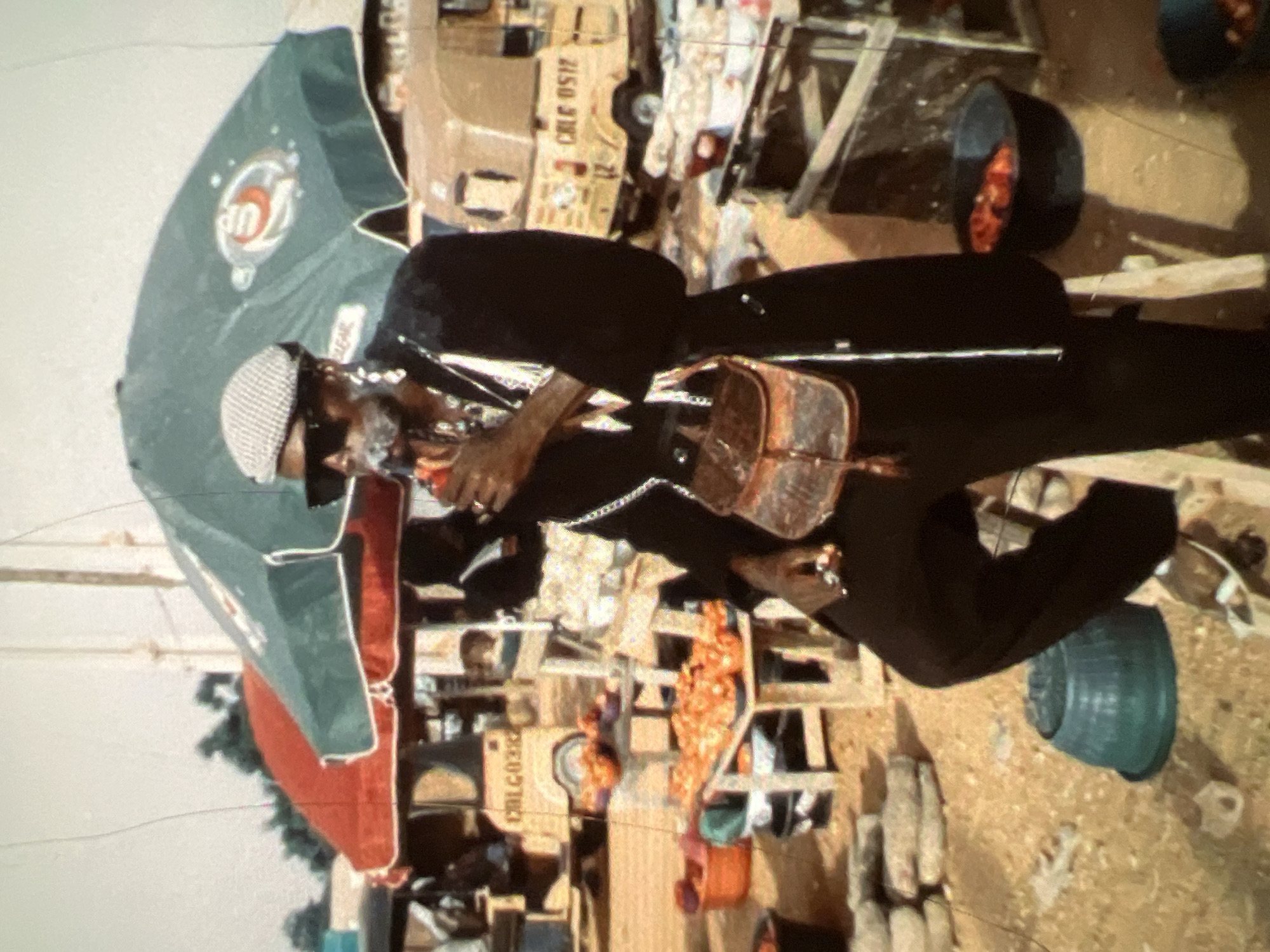 The image depicts a man standing in what appears to be a bustling outdoor market, likely in Lagos, Nigeria, given the presence of distinctive auto-rickshaws, commonly known as 'danfo' buses. The man, wearing a flat cap, sunglasses, a dark coat over a white shirt with a distinctive patterned scarf, and large earrings, is smoking and holding a cigarette. He has a brown leather pouch bag slung across his chest.
The market setting is evident with stalls and a variety of goods. In the background, two auto-rickshaws are visible, one yellow with the license plate "CALG 0517", and another lighter colored one with the license plate "CALG0382". Red and green umbrellas, likely for shade, are prominent over some stalls. Piles of what appear to be tomatoes are displayed on wooden tables, with orange buckets and dark basins containing more produce. The ground is sandy and uneven, typical of an outdoor market. In the foreground, a blue plastic basin sits on the ground. The overall impression is one of vibrant, everyday life in a Nigerian market.