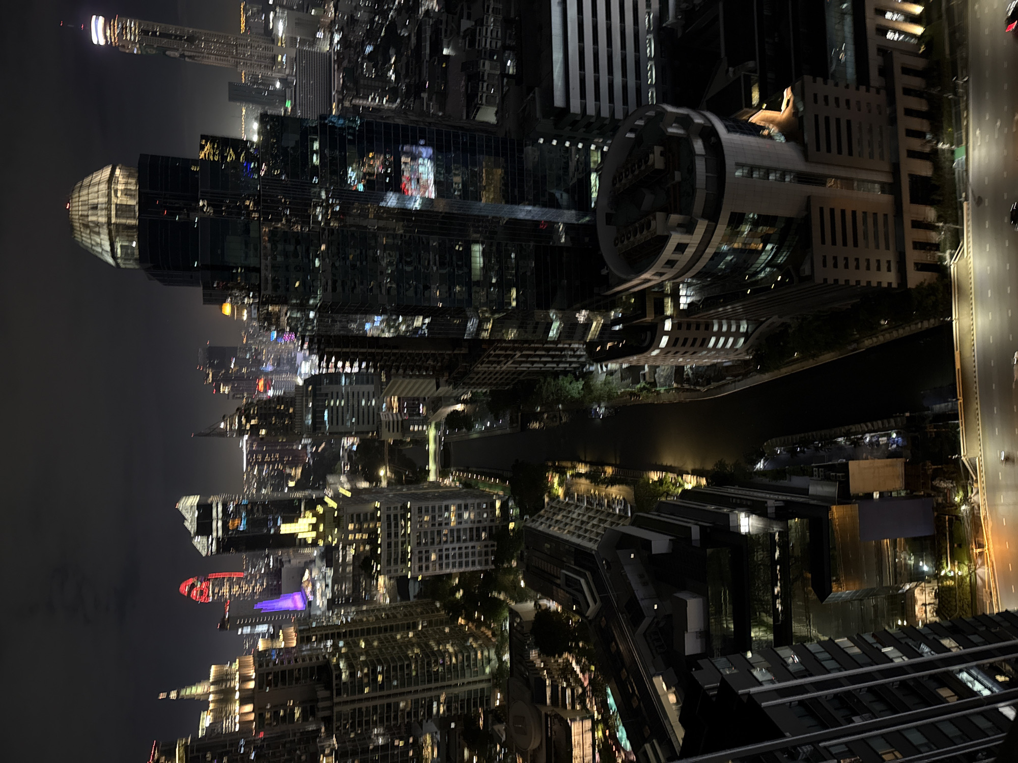 This is a nighttime aerial photograph of a modern city skyline in Bangkok, Thailand, likely taken from a high vantage point such as a skyscraper. The image showcases a dense urban landscape characterized by numerous illuminated high-rise buildings of various architectural designs. A dark, calm river or canal winds its way through the city, reflecting the lights from the surrounding structures. The buildings are a mix of glass-faced towers, some with distinctive geometric shapes and illuminated crowns. One prominent building in the foreground has a multifaceted, domed glass structure at its apex.
The city is aglow with a variety of lights, including white, yellow, and colored illumination, particularly from the windows of residential and commercial buildings, as well as from architectural lighting on the exteriors of some structures. In the distance, taller, more slender skyscrapers are visible, some with illuminated spires. The sky is dark and cloudy, typical of an urban night.
The foreground and midground reveal street-level activity, with cars and their headlights visible on a wide road running parallel to the canal. The scene is one of a bustling metropolis at night, highlighting the impressive scale and luminosity of Bangkok's urban development. There are no discernible people in the image, and the focus is on the architectural grandeur and the urban environment. The overall impression is one of a vibrant and dynamic city.