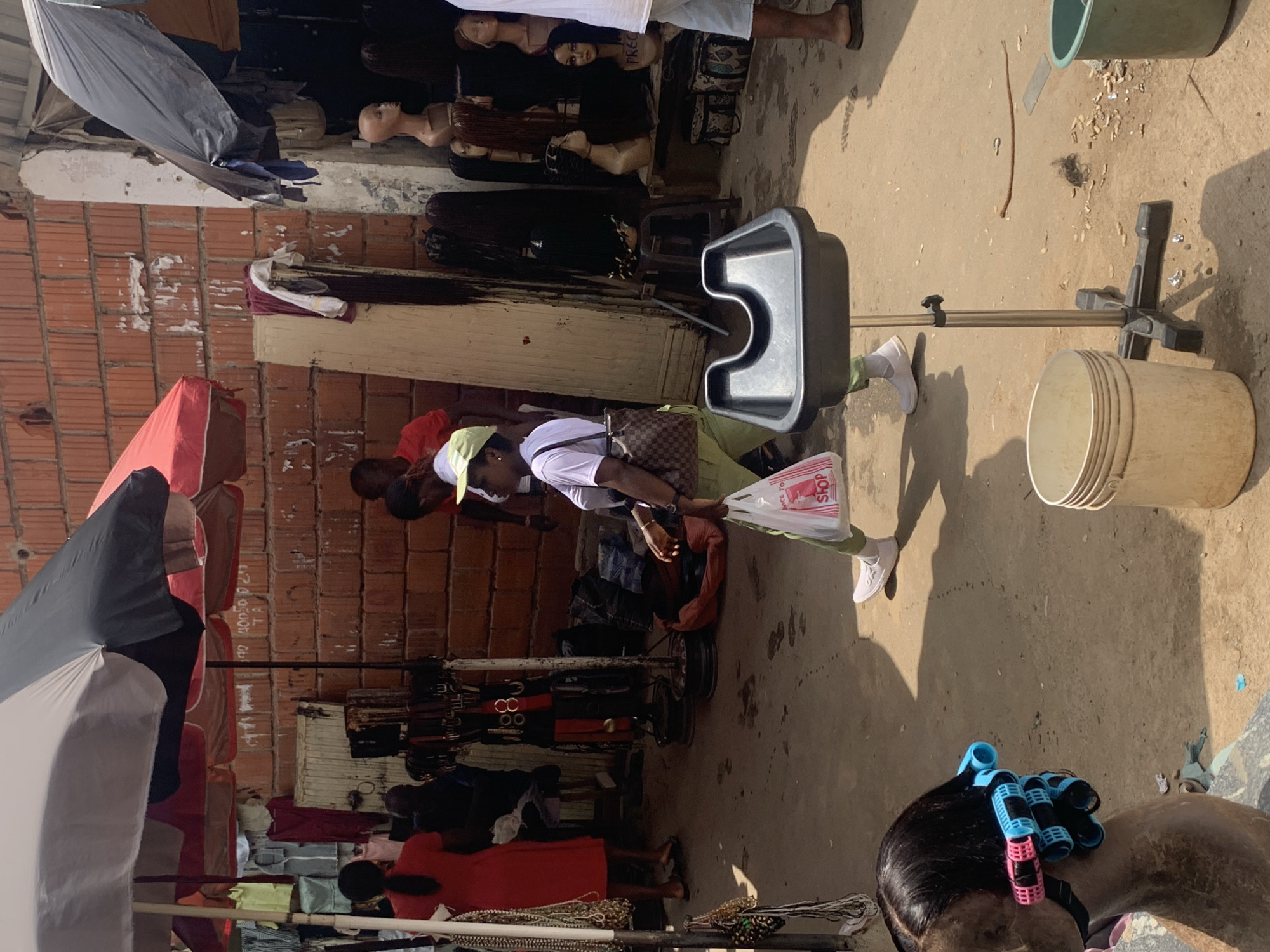 The image depicts an outdoor market scene, likely in Abuja, Nigeria, with various stalls displaying goods under umbrellas. A woman in a white shirt and green pants walks by carrying a shopping bag. In the foreground, a mannequin head with hair curlers is visible. The ground is dusty and appears to be a dirt path. A washing basin on a stand is also present. Brick walls form a backdrop, suggesting the market is set up along a building. Several mannequins displaying wigs are visible in the background. The overall impression is one of a bustling, informal market environment.