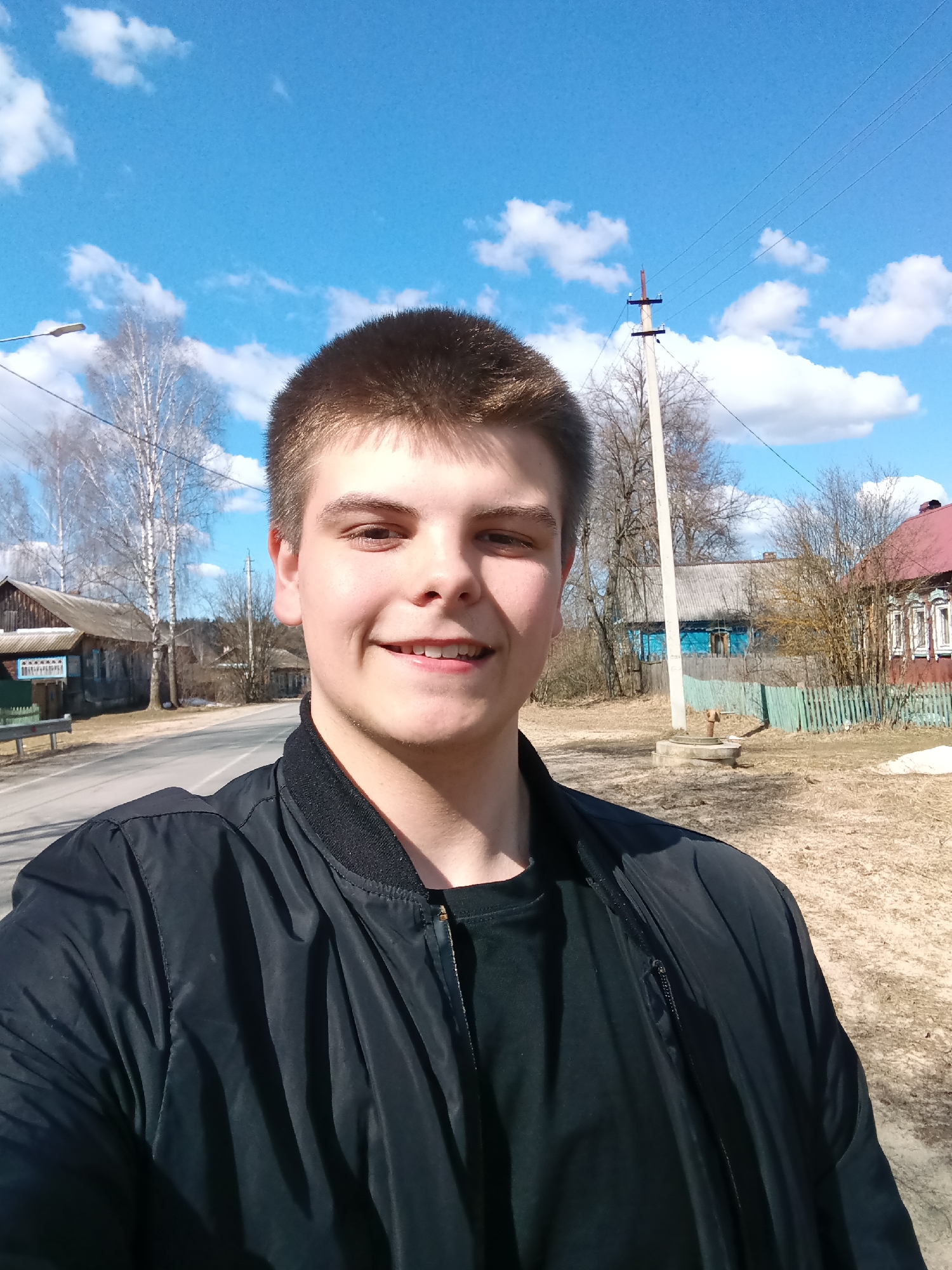 A young man stands in Красный Маяк, Russia, taking a selfie. He is wearing a black t-shirt under a black bomber jacket. Behind him is a paved road, wooden buildings, power lines, trees without leaves, and a blue sky with scattered clouds. The ground is covered with dry vegetation and patches of melting snow.
