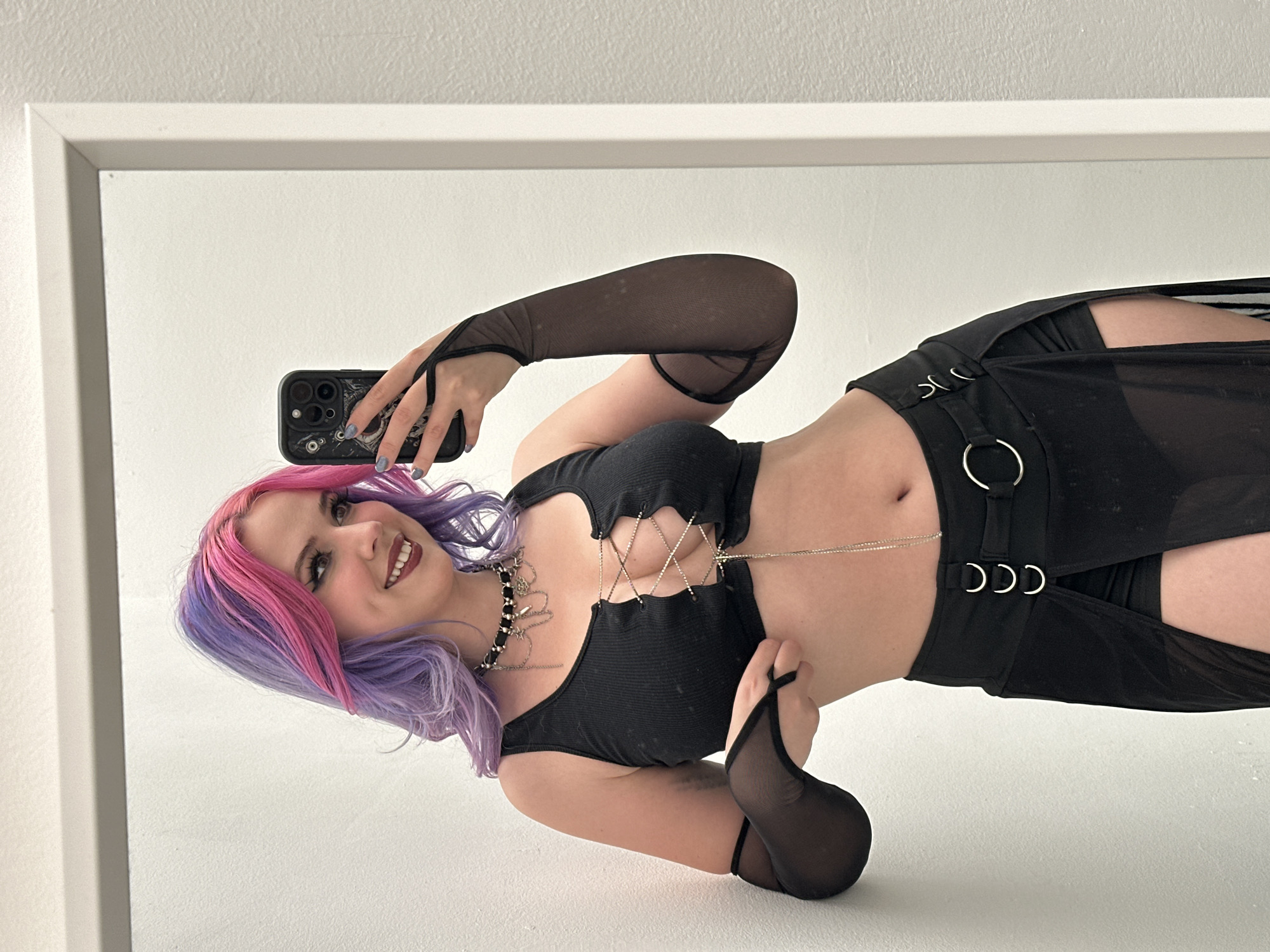 A young woman with pink and purple hair is taking a selfie in a mirror. She is wearing a black top with silver chains and a black skirt with attached rings. She is also wearing black mesh arm sleeves. The background is a plain white wall and a white mirror frame. There is no indication of the location being the City of Zagreb, Croatia.