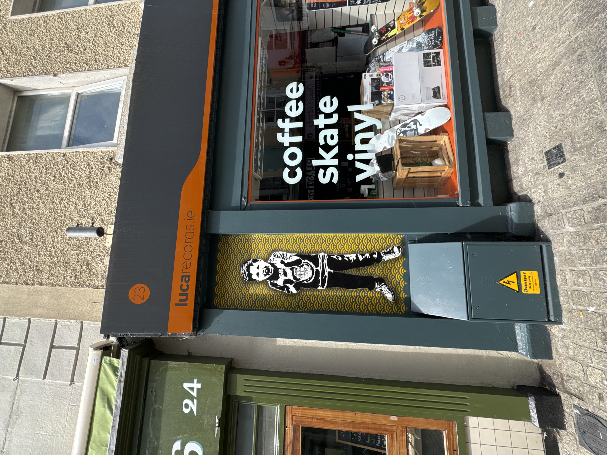 This is a street-level, eye-level, medium shot of a storefront in Waterford, Ireland. The storefront is dark gray with an orange stripe along the top. Above the orange stripe, a circular orange sign with the number 23 is visible. The text "lucarecords.ie" is displayed in white on the orange stripe. To the left of the storefront is a green shop front with the number 24 prominently displayed in white. A large black and white stencil of a young boy is on the dark gray storefront, positioned against a yellow wave pattern. The boy is depicted with his arms raised, giving the middle finger, and wearing a t-shirt with a circular emblem and dark pants. A gray electrical box with a yellow danger sign is situated below the stencil. The shop window displays the words "coffee skate vinyl" in white lettering. Inside the window, skateboards, boxes, and a wooden crate with a decorative item are visible. The ground in front of the storefront is paved with cobblestones.