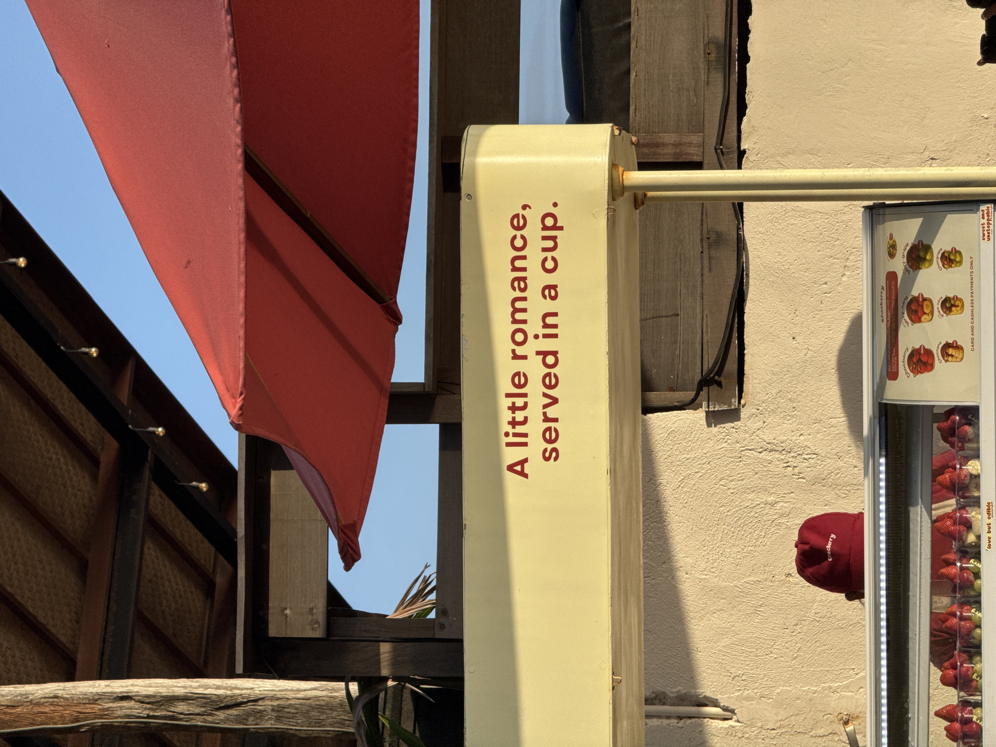 This image depicts an outdoor or semi-outdoor commercial setting, likely a cafe or dessert stall, in North Kuta, Indonesia. The scene is illuminated by bright daylight, casting distinct shadows and suggesting clear, sunny weather.
On the left, a large red fabric awning or umbrella partially obscures the blue sky. To its right, a light yellow vertical beam features prominent red text that reads, "A little romance, served in a cup." A horizontal yellow metal bar extends from this beam across the middle-right of the frame. The background consists of a textured cream-colored wall and dark wooden structures, including beams and a woven roof, adorned with small string lights. Glimpses of tropical foliage are visible through the wooden framework.
On the far right, a vertical display case or refrigerator is partially visible, containing numerous clear plastic cups filled with layered fresh fruits such as strawberries, grapes, and bananas, some appearing to be drizzled with chocolate. Visible text on the display case includes product names like "Chocberry," "Chocnana," and "Chocgrape." Other readable text snippets are "sweet and unstoppable," "love but edible," and "CARD AND CASHLESS PAYMENTS ONLY," indicating payment options. A red cap, possibly part of a uniform or branding, is also seen near the display unit. The overall impression is inviting and suggests a spot offering refreshing treats in a relaxed, possibly tropical, environment.