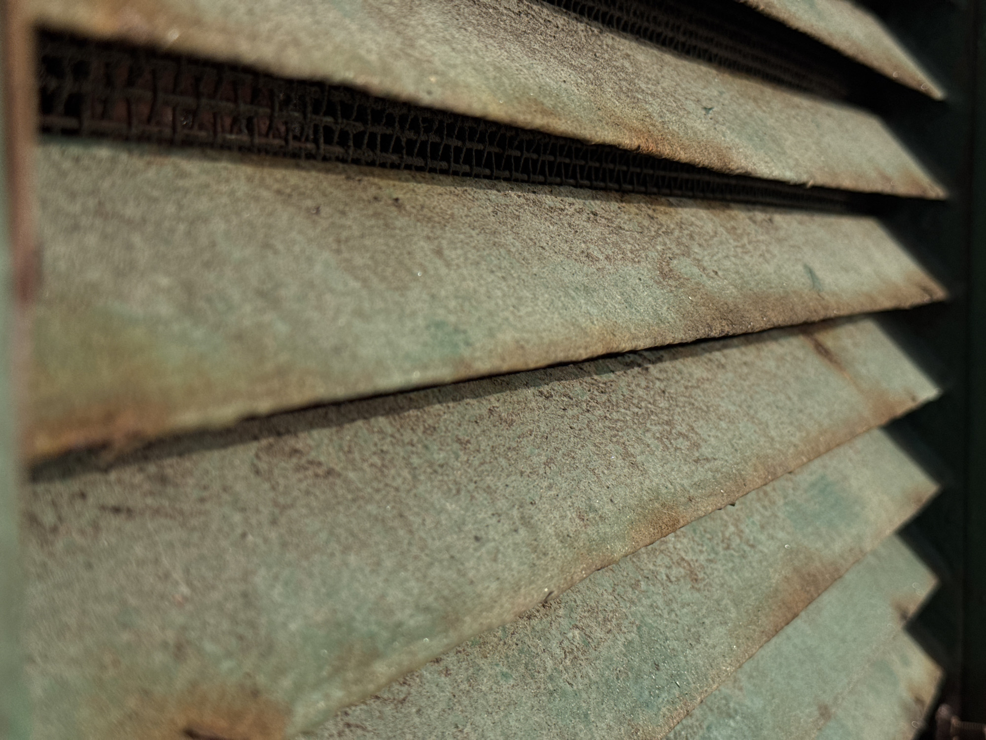 The image is a close-up, shallow depth-of-field shot of weathered, slatted metal. The slats are oriented horizontally and are slightly angled downwards from left to right. They appear to be made of a green-painted metal that is heavily rusted and speckled with dirt or grime. The texture is rough and uneven, with visible pitting and corrosion. Behind the slats, a dark, grid-like structure is visible, suggesting an air vent or grille. The lighting is muted, with shadows cast by the angled slats. The overall impression is one of decay and neglect. The location is not specified in the image, so the city and country remain unknown.