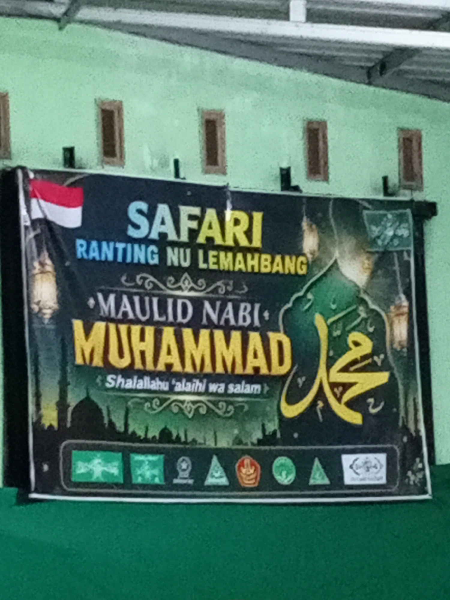 The image shows a banner for "SAFARI RANTING NU LEMAHBANG" which translates to "Safari Branch NU Lemahbang". The banner also features text related to "MAULID NABI MUHAMMAD" which refers to the birthday of Prophet Muhammad. Below this, it says "Shalallahu 'alaihi wa salam," a blessing invoked upon the Prophet. The banner displays several logos at the bottom, including what appears to be the Indonesian flag at the top left. The background of the banner features a silhouette of mosques and minarets, and an artistic rendering of Prophet Muhammad in Arabic calligraphy. The banner is displayed outdoors, with a green surface in the foreground that could be a carpet or fabric. The wall behind the banner is light green and has three rectangular openings. The location is identified as Lemahbang, Indonesia, likely a town or sub-district where a branch of the Nahdlatul Ulama (NU), a major Islamic organization in Indonesia, is located.