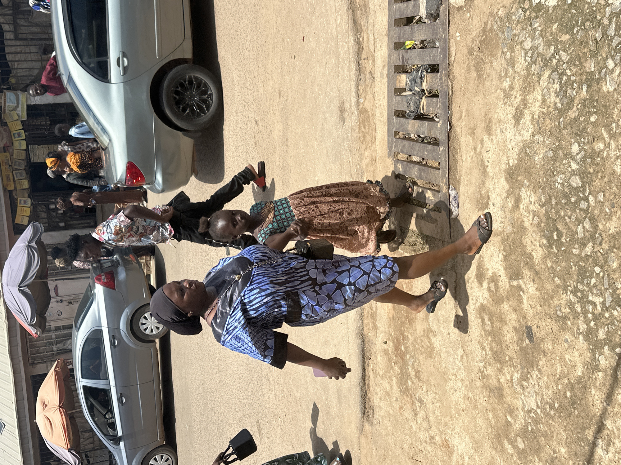 In Narayi, Nigeria, a woman and a young girl walk on a dirt road. The woman, wearing a blue and black patterned dress and a black headscarf, holds a small black purse in her left hand. The girl, in a patterned dress, walks beside her, also holding a black purse. They are walking past a street drain. In the background, a silver sedan is parked, and other pedestrians and buildings are visible, suggesting a busy market area. The sun casts long shadows on the ground.