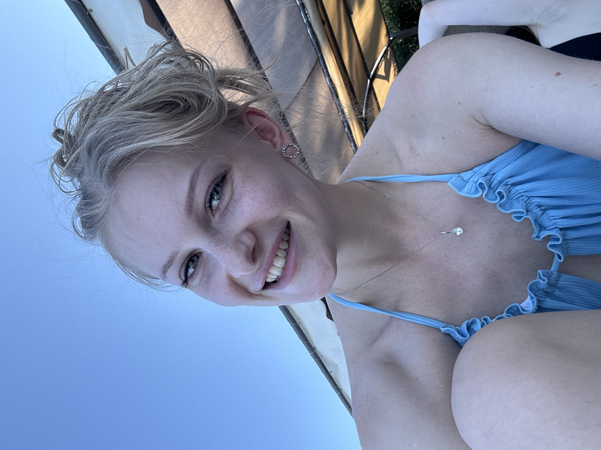 A young Caucasian woman with blonde hair pulled into a messy bun and fair skin with freckles smiles at the camera. She is wearing a blue bikini with ruffled edges and a silver necklace with a teardrop-shaped pendant. She is also wearing circular earrings with small stones. The background shows a clear blue sky and part of an outdoor canopy structure. The overall scene suggests a relaxed, outdoor setting, possibly a vacation or a day at the beach in Kemer, Turkey, given the blue sky and coastal ambiance.