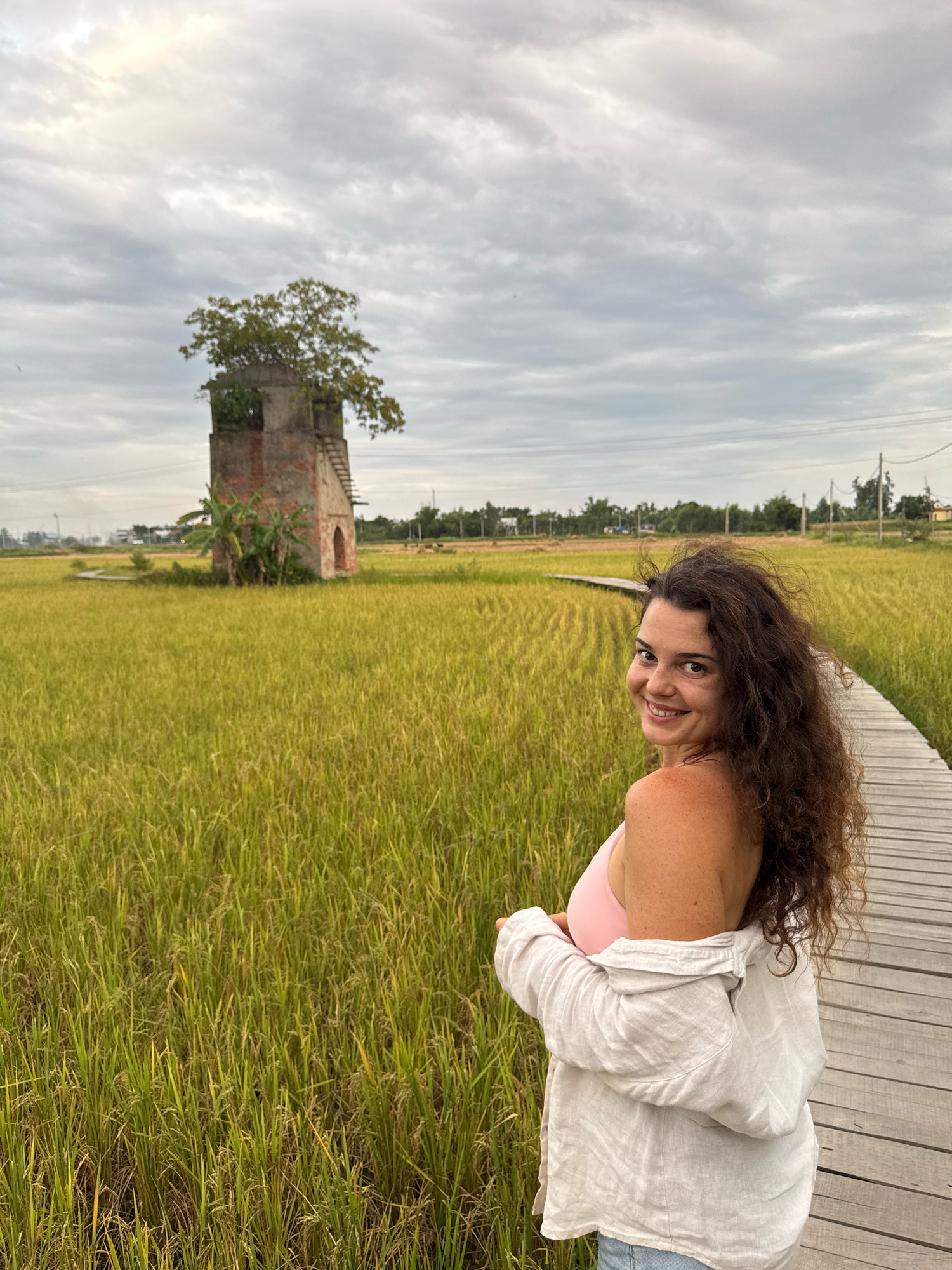 In Đà Nẵng, Vietnam, a woman with long, curly brown hair is captured in a portrait with a wooden boardwalk curving through a lush, green rice field. She is looking over her shoulder and smiling at the camera, her face illuminated by soft daylight. She is wearing a light pink bikini top and an unbuttoned white linen shirt. In the background, a weathered brick structure with a tree growing from its top stands amidst the rice paddies, under a cloudy sky. The scene is tranquil and evokes a sense of rural beauty.