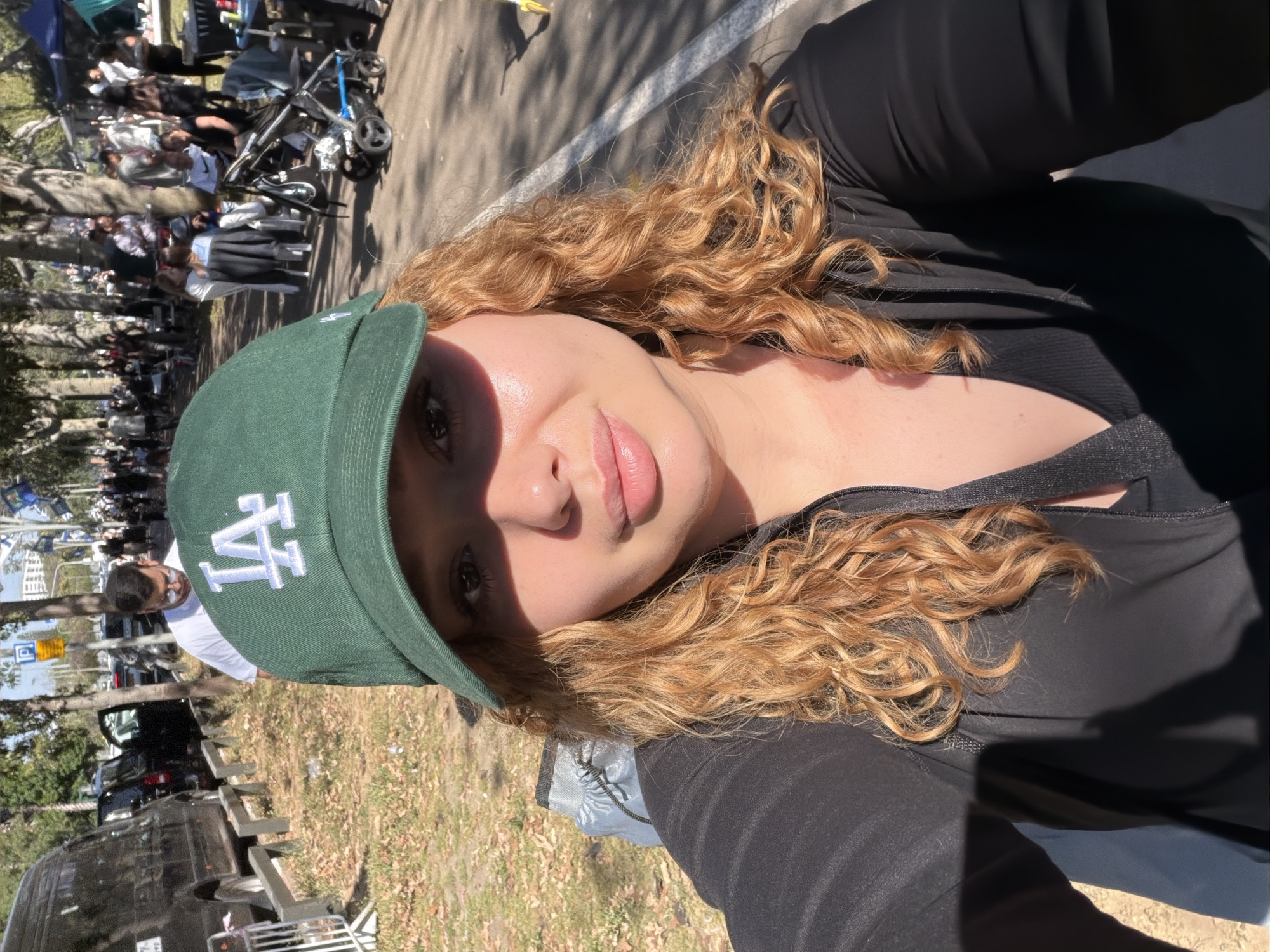 The image is a selfie of a young woman, presented rotated 90 degrees clockwise. She dominates the foreground, with a bustling outdoor scene behind her.
**Subjects:**
The primary subject is a woman with long, wavy, light brown or auburn hair. She wears a dark green baseball cap featuring a prominent white "LA" logo on the front. Her face, partially shadowed by the cap and sunlight, reveals her eyes and full, pink lips. She is dressed in a black long-sleeved top or jacket, possibly activewear, with a dark strap visible near her shoulder.
In the background, a large crowd of people is visible, dispersed along a paved path or road bordered by trees. Individuals of various ages, including men, women, and children, can be seen, with some pushing baby strollers. In the distance, several vehicles, including a dark van or SUV, are parked or moving. The ground features patches of dry grass and dirt.
**Activity or event taking place:**
The gathering of numerous people, strollers, and vehicles suggests a public event, a busy day in a park, or a local market or festival. The atmosphere appears casual and social.
**Notable details:**
The scene is brightly illuminated by natural sunlight, casting distinct shadows, indicating it is daytime, likely midday or early afternoon. The weather appears clear and sunny. A blue street sign with a white "P" (parking) symbol is faintly visible in the background. The "LA" logo on the cap, often associated with the Los Angeles Dodgers, is a clear visual detail. The overall setting implies a public space in an urban or suburban environment, aligning with the provided context of Ramat Gan, Israel.