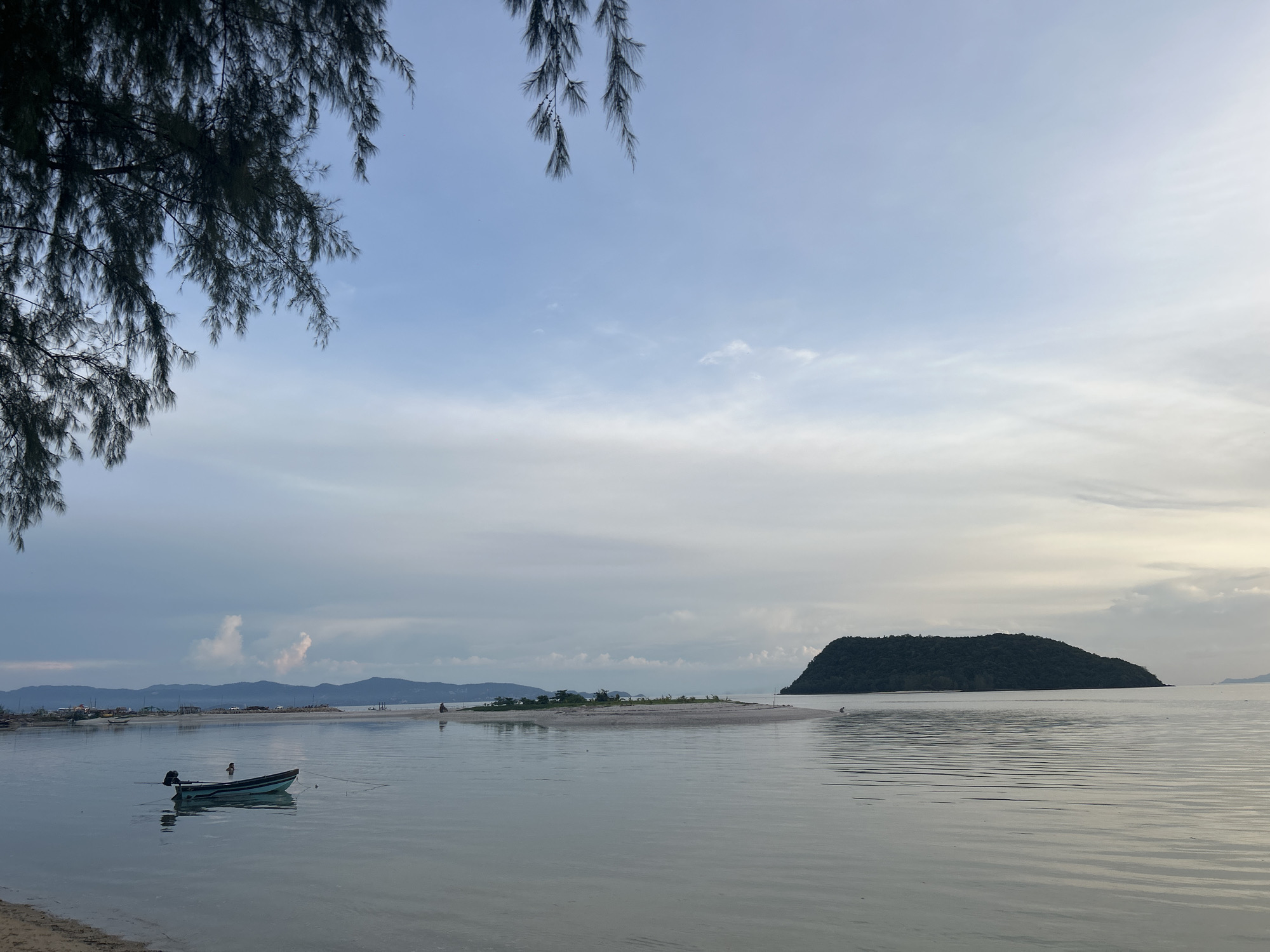 This image depicts a serene coastal scene in ตำบลเกาะพะงัน, Thailand, likely at dawn or dusk given the soft lighting. In the foreground, the calm, reflective water meets a sandy shore partially visible at the bottom left. A small, dark-colored boat with a turquoise trim is anchored in the shallow water, its reflection slightly distorted on the surface. A person is visible standing in the water near the boat. Further out, a thin strip of sand forms a sandbar, with several small trees dotting its surface. In the mid-ground, a larger, heavily forested island with a distinct silhouette rises from the sea. The horizon is marked by a range of distant hills or mountains, possibly part of the mainland, with buildings and lights suggesting a settlement. The sky is a gradient of pale blue and white, with scattered clouds, hinting at an overcast or transitional weather pattern. A few branches of dark, needle-like foliage hang into the frame from the top left, suggesting the presence of pine or similar trees at the edge of the viewpoint.