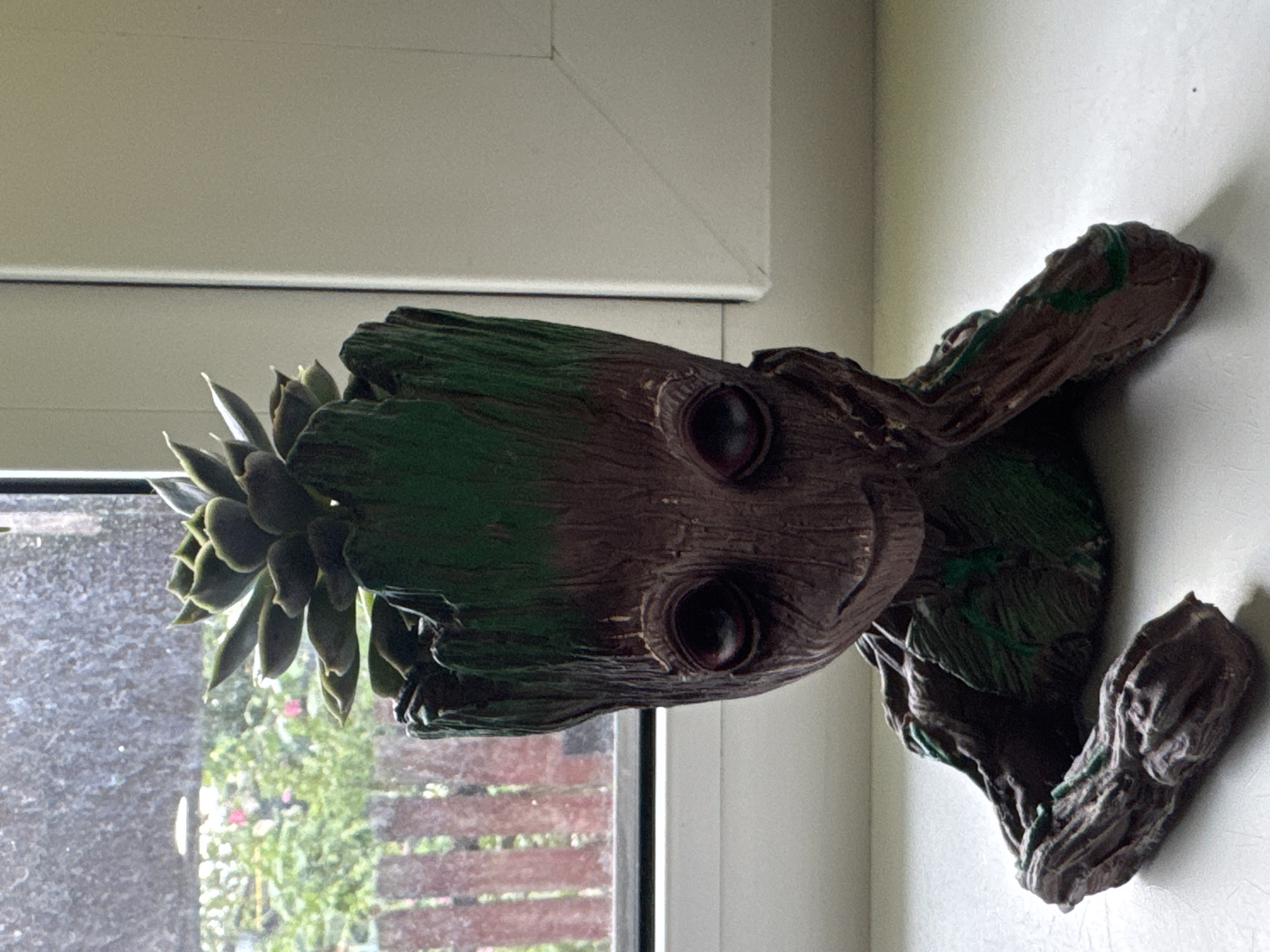 The image shows a Groot-shaped planter containing a succulent plant. The planter is brown with green accents and has a distressed, wood-like texture. The succulent has thick, fleshy green leaves. The planter is positioned on a white windowsill. Behind the planter, a window looks out onto a backyard with a wooden fence and some foliage. The overall lighting suggests it is daytime, possibly overcast. The image was taken in Kremenchuk, Ukraine.