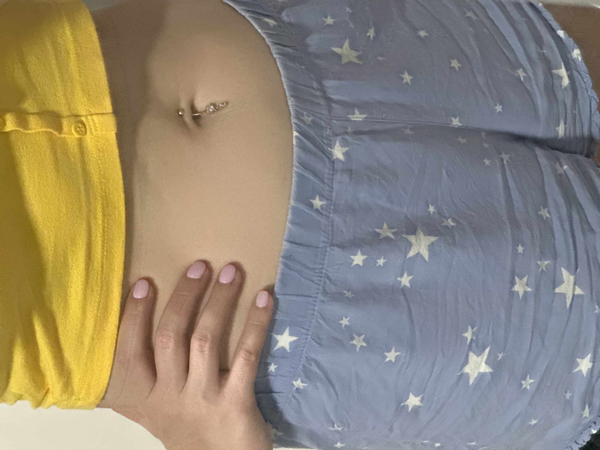 The image shows a close-up view of a person's midsection, specifically the stomach area. The person is wearing a yellow crop top and light blue shorts adorned with white stars. A navel piercing with a dangling charm is visible. A hand with light pink painted fingernails rests on the person's hip. The background is a plain, light-colored wall. The image does not provide any information about the location in the city of Nevinnomyssk, Russia.
