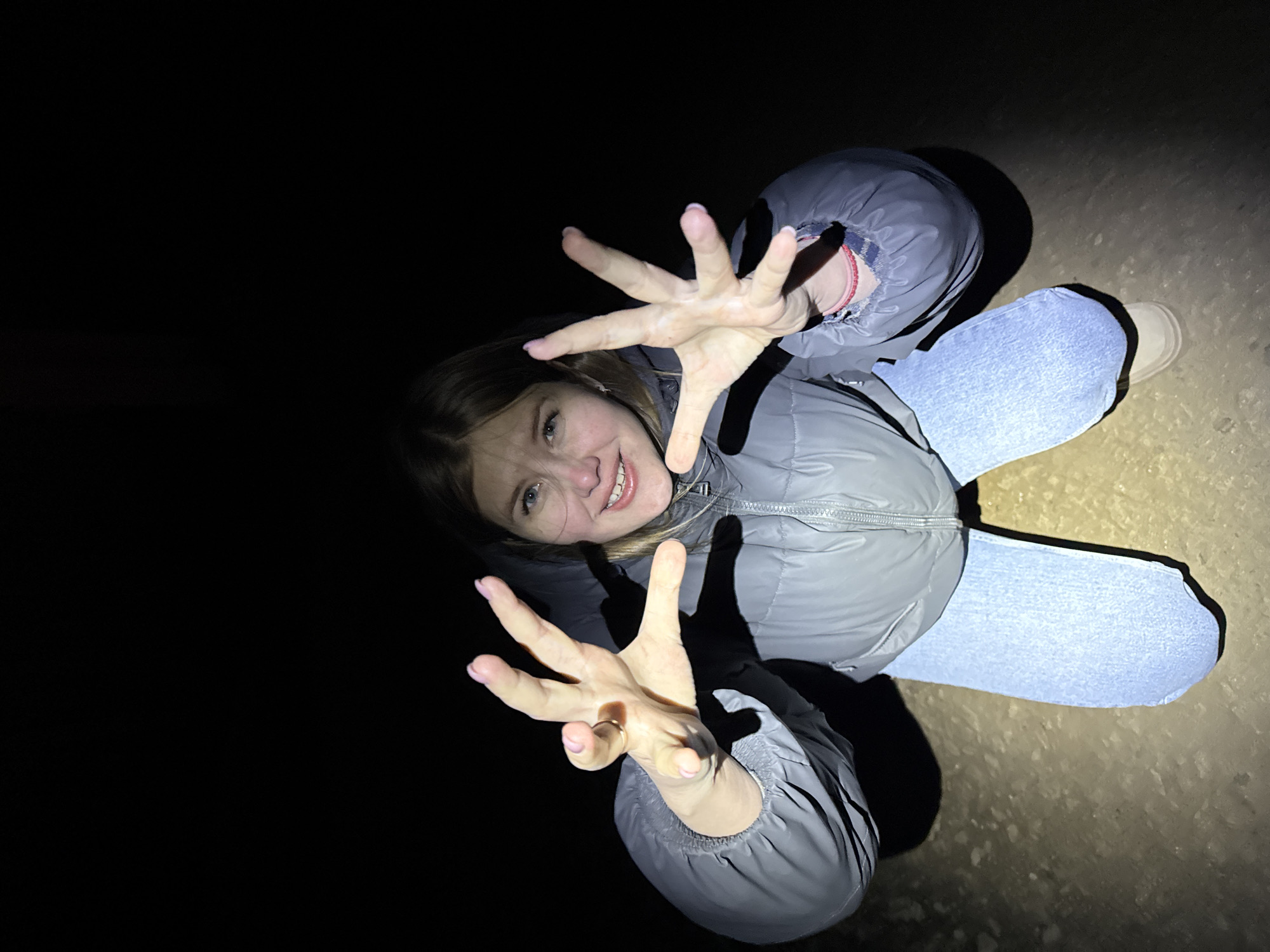 A young woman is captured in a high-angle shot, lying on her back on a textured ground surface, and looking up towards the camera. The scene is enveloped in darkness, suggesting it's nighttime or a very dimly lit environment, with only the subject and the immediate area around her illuminated by a strong, direct light source, likely a flash or flashlight. This harsh lighting casts prominent shadows behind her body and arms onto the ground.
The woman is smiling broadly, revealing her teeth, and appears cheerful and playful. She extends both of her hands towards the camera, fingers spread wide open, as if reaching out or making a playful gesture. She is wearing a light grey puffy jacket with a zippered front and light blue jeans. Her fingernails are painted with light pink polish. On her left hand (viewer's right), a gold-colored ring is visible on her ring finger, and on her right wrist (viewer's left), a red and white braided bracelet can be seen.
The ground beneath her appears to be a rough, possibly gravelly or sandy, light-colored surface. The background beyond the illuminated area is completely black. The overall impression is a candid, informal portrait taken in the dark, with the subject engaging directly with the camera. The image was taken in Горный, Russia.