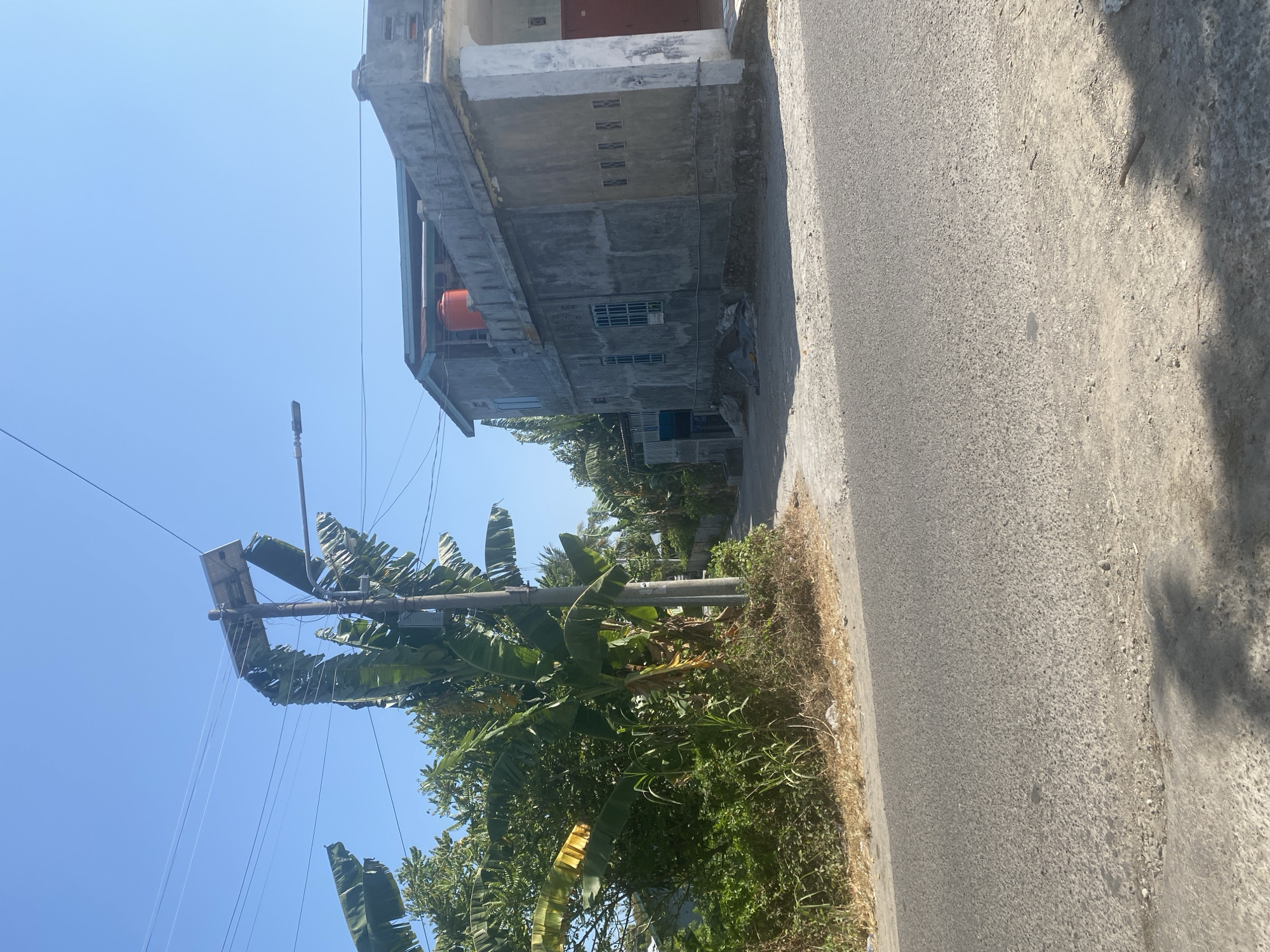 This is a daytime, eye-level, outdoor, medium shot capturing a scene on a paved road in the city of Mattekko, Indonesia. The road curves slightly to the left in the foreground, leading towards a junction. On the right side of the road, a concrete building with a grey, unfinished facade stands. The building features several small, barred windows and a prominent orange water tank on its roof. An arched entryway with a dark red door is visible on the right side of the building. Adjacent to the building, a narrow alley or path recedes into the background. On the left side of the road, dense vegetation, including banana trees with large leaves and other green foliage, lines the roadside. A tall utility pole stands amidst the vegetation, equipped with a solar panel at its top and a street light attached to its side. Power lines stretch across the upper portion of the image. The sky is clear and bright blue. The ground along the roadside is covered with dry leaves and debris. There are no people or animals visible in the frame.