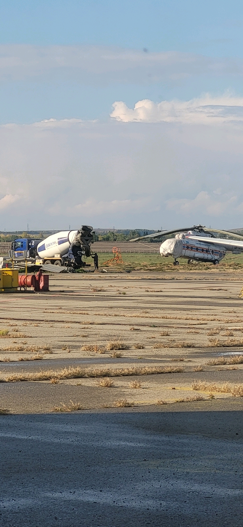 A concrete mixer truck and a helicopter are parked on an airfield. The concrete mixer truck is blue and white, with "SKAMEL" written on the side of the drum. The helicopter is white with red and blue stripes. It is covered with a tarp. There are a few people standing near the concrete mixer truck. The ground is cracked concrete with dry grass growing through it. The sky is blue with some clouds.