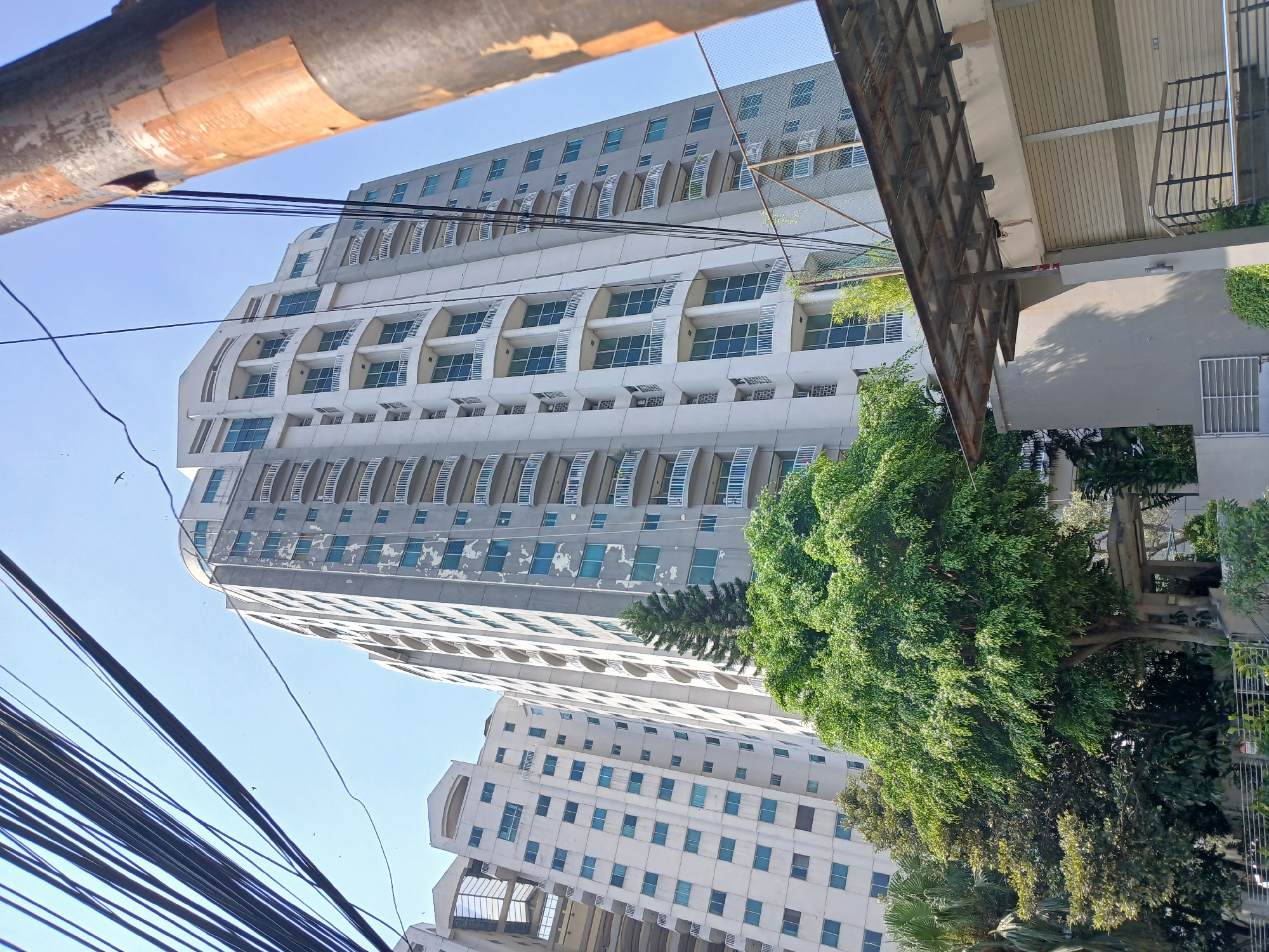 The image depicts a cluster of tall residential buildings against a clear blue sky. The building in the foreground, on the right, is a light grey skyscraper with numerous balconies and light blue windows. Its facade shows signs of wear, with peeling paint in several areas. To the left and slightly behind this building is another, taller skyscraper, also predominantly light grey with balconies and blue windows. Further to the left, another, less detailed building with a more geometric facade and rows of windows is visible. Lush green trees and foliage are prominent in the foreground, partially obscuring the lower levels of the buildings. A thick bundle of black electrical wires crisscrosses the upper portion of the image, and a rusty metal pole is visible on the far right. A low-lying structure with a metal roof and roller shutters is in the lower right quadrant. There are no people visible in the image. The scene appears to be an urban environment in South Jakarta, Indonesia, likely a residential complex.
