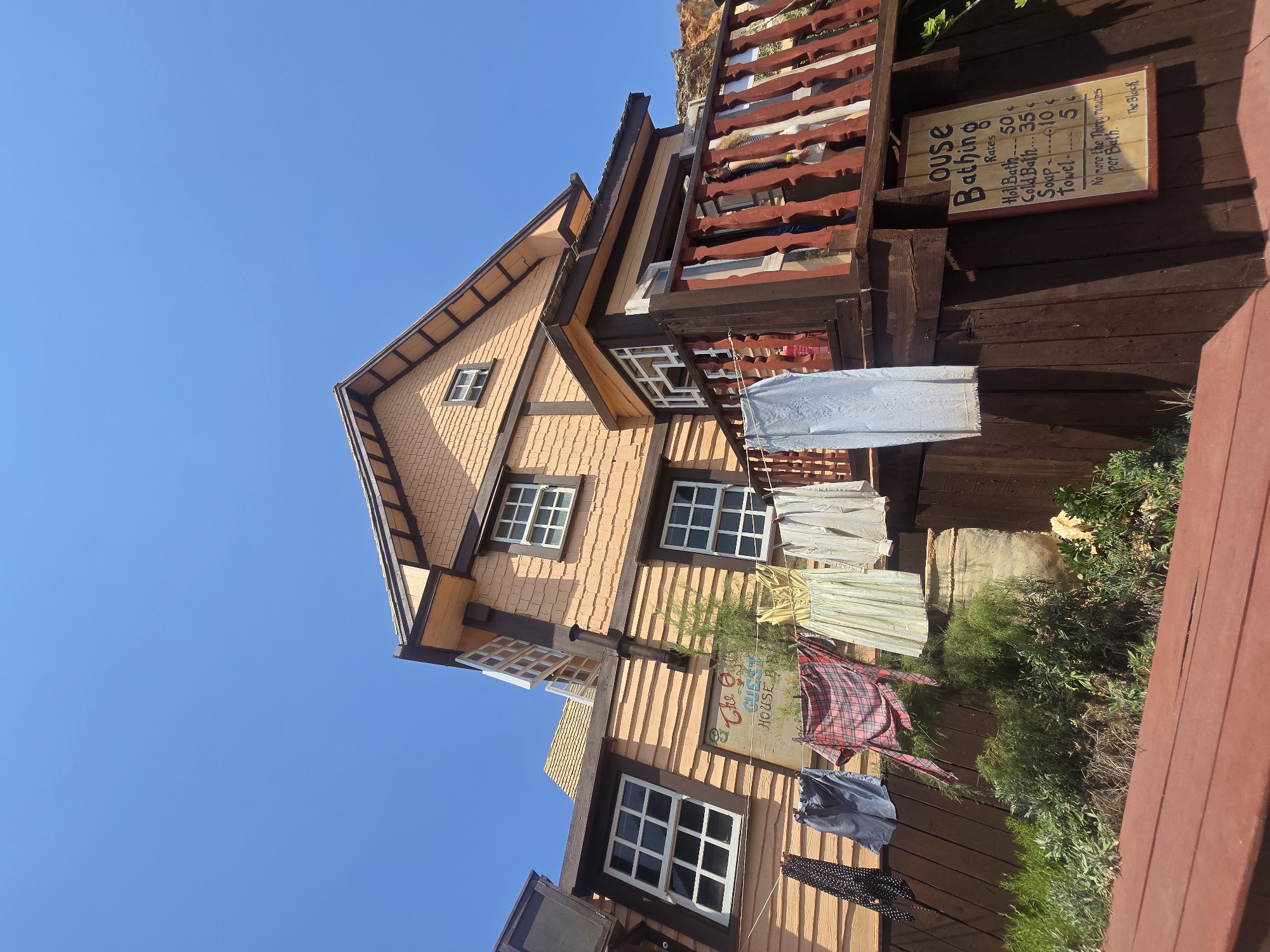 The image depicts a quaint, rustic building exterior, likely a guesthouse, set against a clear blue sky. The building features an orange-toned wooden facade with visible horizontal siding. Several windows with white frames and multiple panes are visible on the upper floors. Laundry, including a black and white polka-dot garment, blue trousers, a red and black plaid shirt or dress, a yellow dress, and white shirts, hangs on a clothesline strung across the front of the building.
Below the clothesline, a sign with red lettering on a cream background reads "The Ostrich Guest House". Further down, a second, more detailed sign attached to the wooden structure displays "Ouse Bathing Rates" with a list of services and prices: "Hot Bath 50¢", "Cold Bath 35¢", "Soap 10¢", and "Towel 5¢". Below this, a handwritten note states, "No more than Thirty Minutes per Bath. The Black".
The building has a wooden balcony with red-painted railings. Green foliage and bushes grow around the base of the building, and a large, rustic ceramic pot is partially visible among the greenery. The overall aesthetic suggests a charming, possibly historic, tourist attraction. This scene is consistent with the appearance of the Popeye Village in Mellieħa, Malta, which is a purpose-built film set that has been preserved as a tourist attraction and resembles a quaint, old-fashioned village.