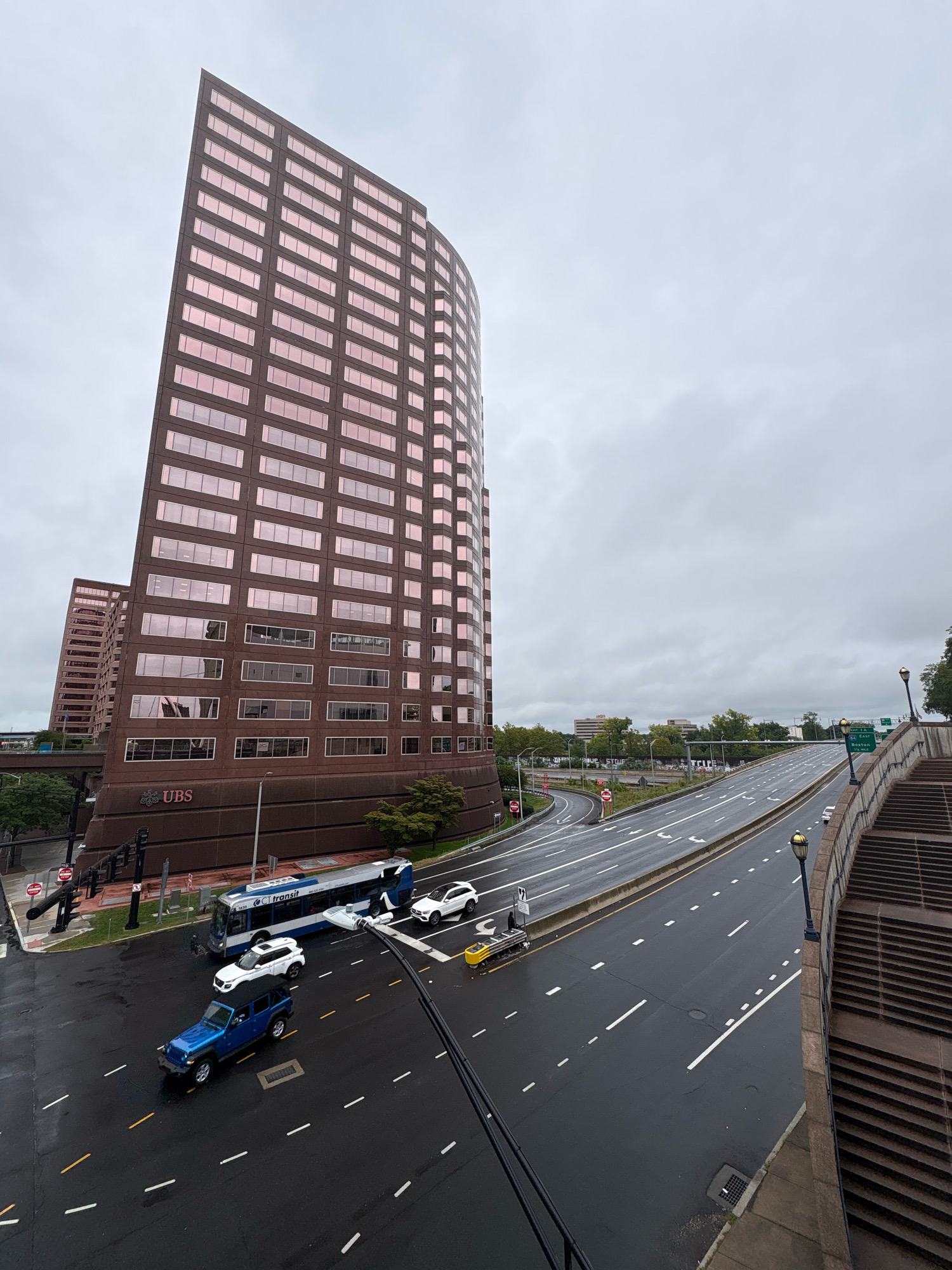 This image captures a street scene in Hartford, Connecticut, USA, dominated by a large, angular, rose-colored glass-and-steel office building. The building has a distinctive curved facade and numerous windows reflecting the overcast sky. In front of the building, a multi-lane road curves into the distance. A CT transit bus and several cars, including a blue Jeep, a white SUV, and a white sedan, are visible on the wet road, suggesting recent rain. A yellow construction vehicle is parked on the side of the road. On the right side of the image, a concrete staircase leads upwards, bordering the road. Traffic lights and street lamps are present, and a highway exit sign pointing towards Boston is visible in the background. The sky is a uniform gray, indicating cloudy weather. The overall impression is an urban environment on a damp, overcast day.