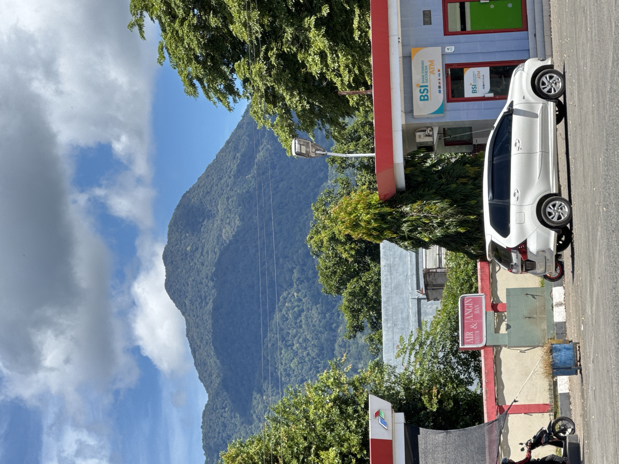 The image depicts a scene in Sukamulia, Indonesia, with a mountainous backdrop. In the foreground, a white van is parked in front of a building with a blue facade and red trim. The building has signage for "BSI BANK SYARIAH INDONESIA ATM." To the left of the van, there is a gas station canopy with red and white detailing and a red sign that reads "AIR & ANGIN." A person on a motorcycle is pulling away from the gas pump area. Behind the gas station, there's a wall and a cluster of trees. The mountain in the background is covered in lush green vegetation, and the sky is a clear blue with scattered white clouds. The overall impression is a typical day at a service station in a tropical or subtropical region, with a prominent natural landscape feature.