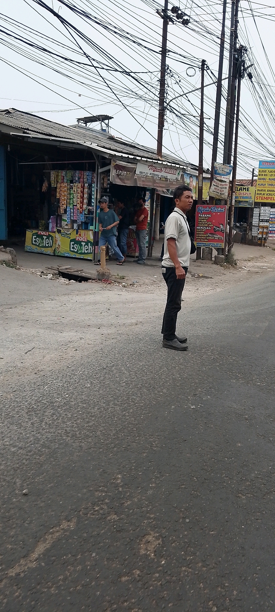 This image depicts an outdoor scene on a street in Cileungsi, Indonesia, characterized by a low-lying building with a corrugated metal roof, likely a shop or stall, and a dense network of overhead electrical wires. A man wearing a light-colored short-sleeved shirt, dark pants, and dark shoes stands in the foreground on the right, facing to the right with his body angled towards the viewer. He has a bag slung over his left shoulder. Behind him, to the left, several other individuals are visible near the building. One person in a blue t-shirt and blue jeans is walking. Another person in a red shirt and dark pants is standing. The building appears to be a retail establishment, with various products displayed on shelves and signage visible, including advertisements for "Eso Teh" and "Mando Variasi". The ground is paved with asphalt, and there is evidence of disrepair with a visible drain or ditch in the road. The overall environment suggests a commercial area with significant overhead infrastructure.