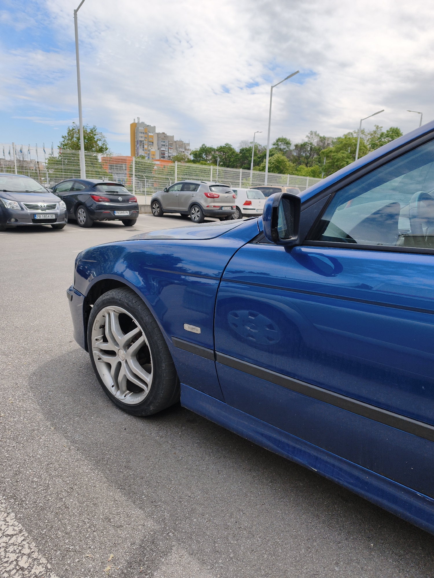 A blue car is parked in a parking lot. In the background are other cars including a gray Toyota Avensis with license plate PB 1854 XE, a dark gray Opel Astra with license plate PB 5445 PK, and a gray Kia Sportage. A white fence separates the parking lot from a residential area with apartment buildings. The parking lot has street lights. The location is Plovdiv, Bulgaria.