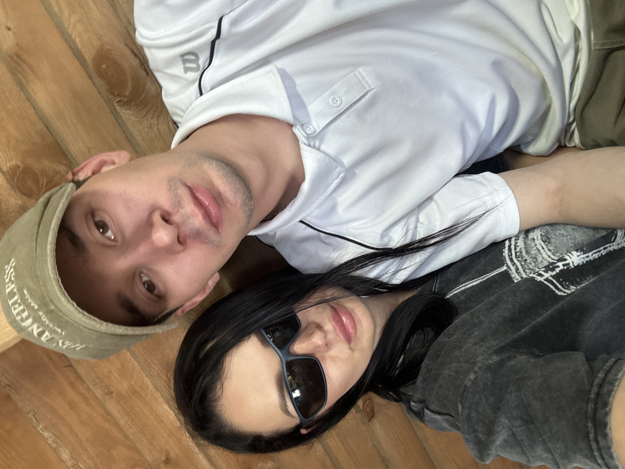 In this close-up, selfie-style image, a couple is depicted against a backdrop of rustic wooden walls. The woman, on the left, has long, dark hair and is wearing dark sunglasses and a distressed grey t-shirt. The man, on the right, is wearing a green baseball cap with "LOS ANGELES" embroidered on it, and a white polo shirt with a black trim. His face is visible from the nose up, and his right shoulder is slightly turned towards the camera. The setting appears to be indoors, possibly a cabin or a room with wooden paneling, suggesting a location in Votkinsk, Russia, given the context. The lighting is natural, casting soft shadows on their faces.