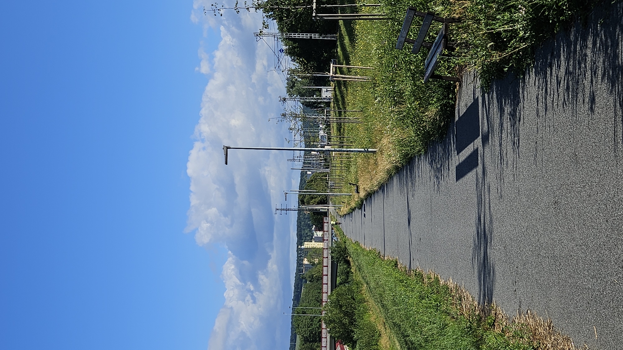 An asphalt path runs through a park area adjacent to a grassy embankment on the left and a grassy area with young trees and streetlights on the right. The path recedes into the distance where a bridge crosses a body of water. A single person is visible cycling on the path, and a park bench is situated on the right side of the path. In the background, there are buildings and electrical infrastructure, including power lines and towers. The sky is blue with scattered white clouds. The scene appears to be in Prešov, Slovakia, along the Torysa river, based on the presence of the river and the typical urban park and infrastructure seen in the city.