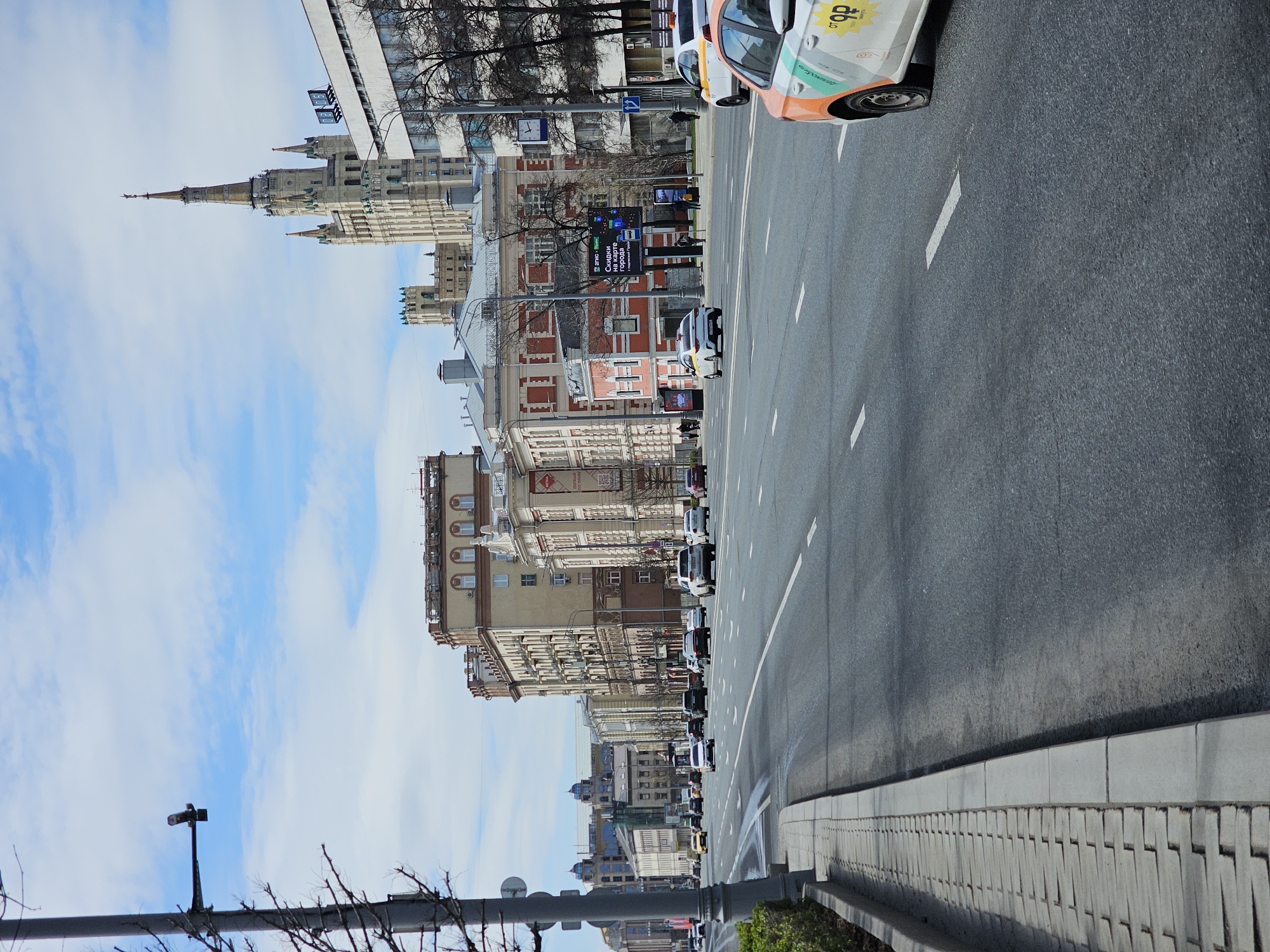 The image depicts a street scene in Moscow, Russia. A wide asphalt road occupies the right half of the frame, marked with white lane dividers, and a car partially visible in the upper right. A pedestrian sidewalk runs along the road's edge. A line of various architectural buildings lines the left side, ranging in color, height, and style. The background features a Stalinist skyscraper in a lighter color. The sky above is blue, dotted with light clouds. Several cars are visible on the road.