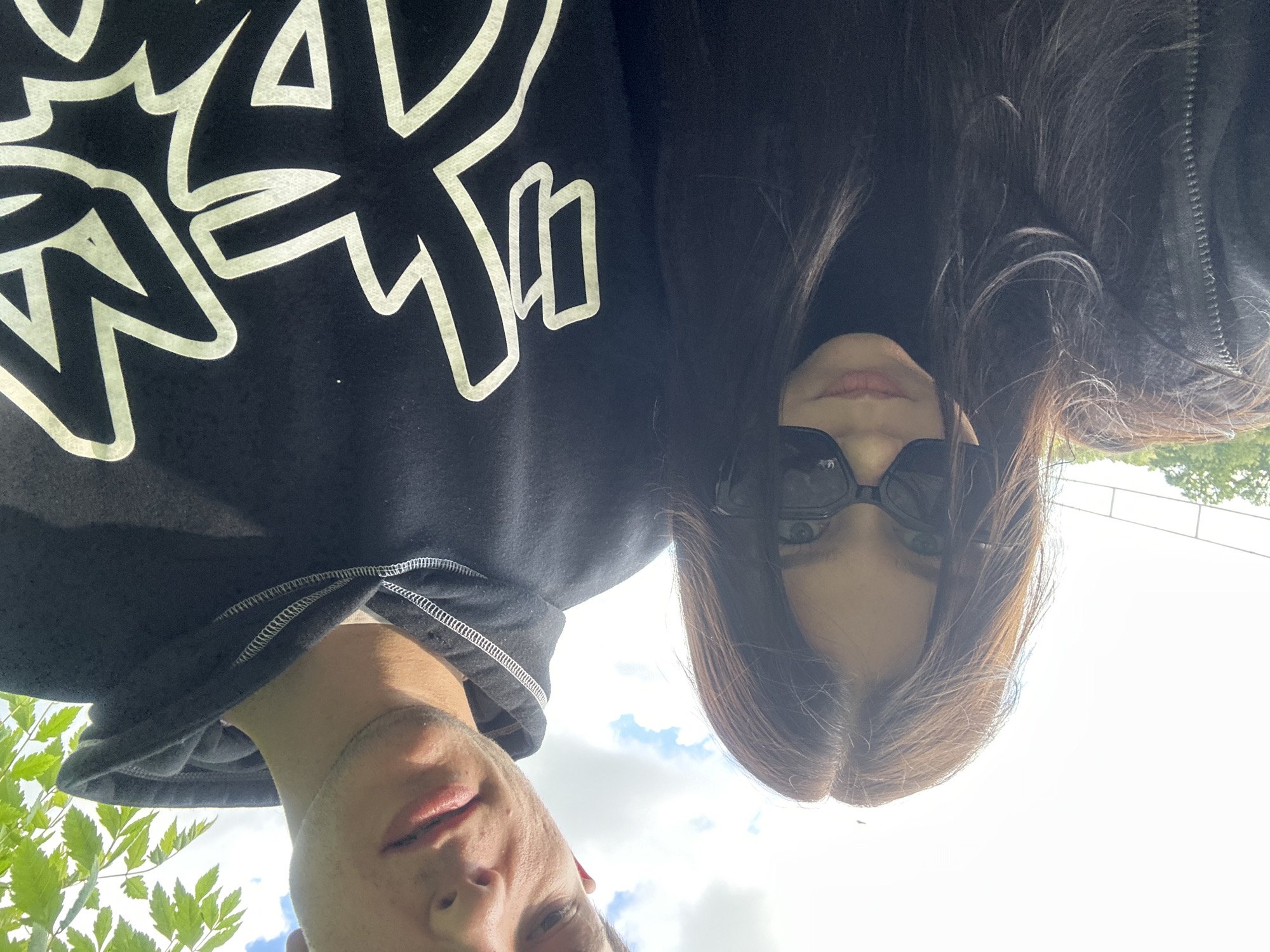 A low-angle shot depicts two individuals outdoors in Beloslav, Bulgaria. The camera is tilted, presenting a skewed perspective of the subjects and environment.
Person 1, a male, occupies the left side of the frame. He is wearing a black hooded sweatshirt with a large, white graphic design on the front. His face is partially visible, and some green foliage is visible near his neck and shoulder.
Person 2, a female, appears to the right, wearing sunglasses. Her dark hair frames her face. The sky with some clouds are visible in the background. The angle suggests the photo was taken by someone holding the camera low and angled upwards.
