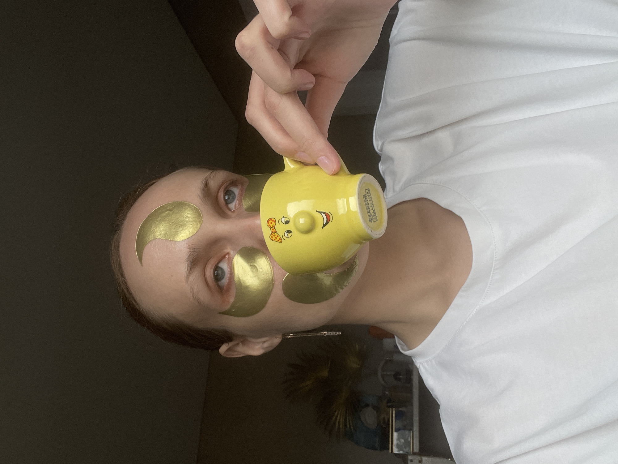A woman with light skin and light brown hair is angled to the left of the frame, wearing gold crescent-shaped cosmetic patches under and above her eyes. She is wearing a white t-shirt. Her right hand is holding a yellow ceramic mug with a cartoon face on it. The mug obscures her face, specifically her nose and the middle section of her forehead. The handle of the mug is oriented upward. Behind her is a dimly lit room with shelves and a plant. There's no indication of the location being Podshivalovo, Russia.