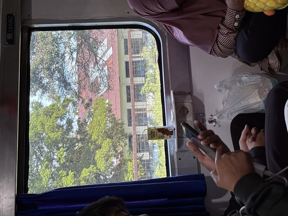 Inside a train car, passengers are seated with a view through the window revealing trees and a building. An electrical outlet is visible near the window, along with a small carton of Ultra Milk. A person holds a smartphone, and the blue curtain lining the bottom of the window is partially visible. The scene is captured in natural light.