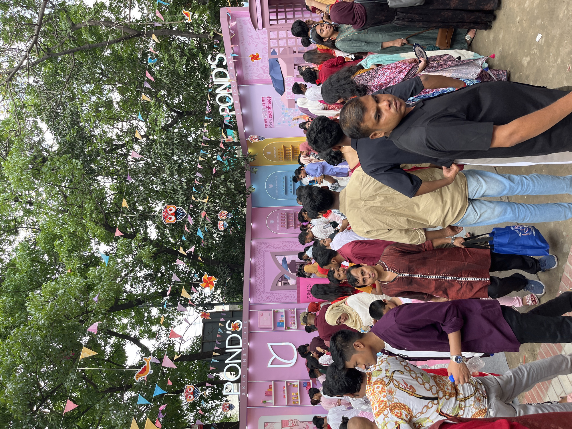 The image shows a large gathering of people in Dhaka, Bangladesh, at what appears to be a promotional event for the skincare brand POND'S. A pink backdrop featuring the POND'S logo and advertisements for its products is visible. Above the crowd, colorful pennants and decorations are strung from trees. The people in attendance are of various ages and genders, dressed in everyday clothing, and seemingly interacting with the event or each other.