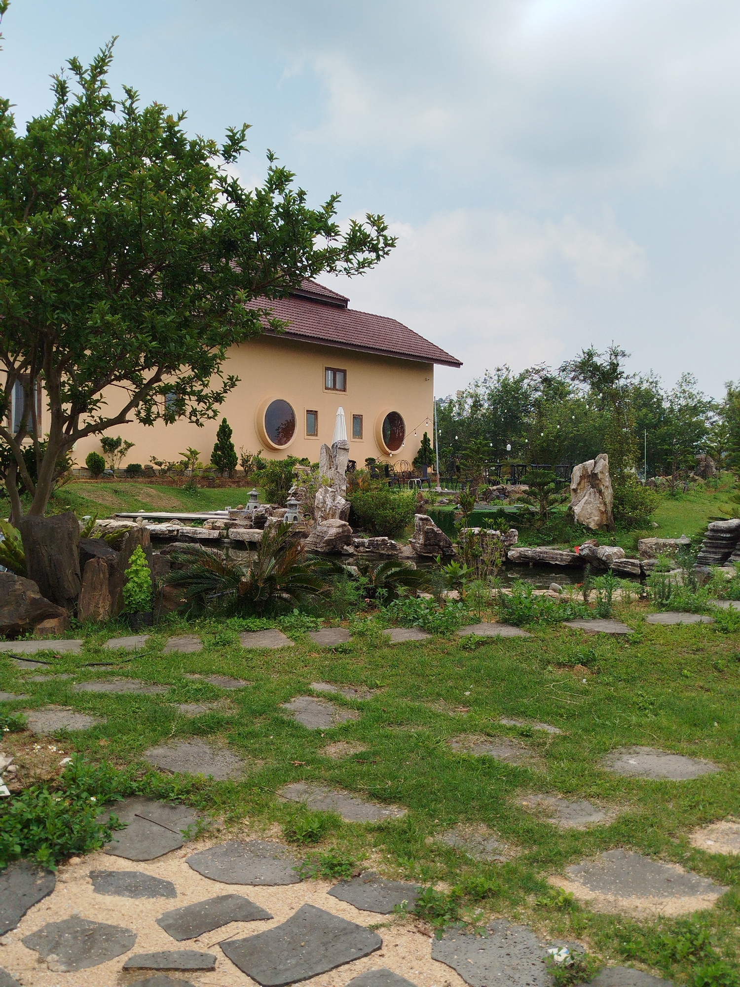 The scene is a garden in Đa Nghịt 1, Vietnam, with a unique building in the background. The building has a tan facade, a brown tiled roof, and a mix of square and circular windows. A large tree stands to the left of the building, partially obscuring its facade.
The foreground shows a stone pathway leading towards a water feature and garden area. This area is adorned with various rocks, plants, and small decorative stone lanterns. The lighting suggests an overcast day. Further back, there is more greenery and some tables and chairs under string lights.