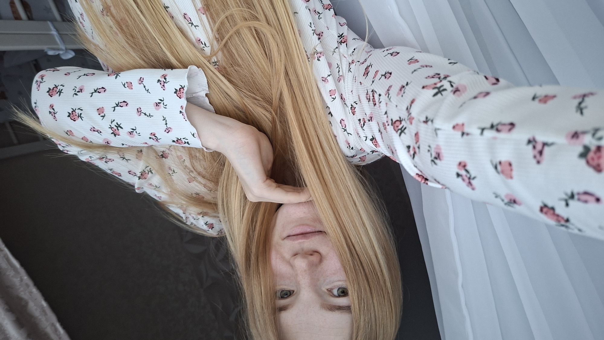 The image is a close-up selfie featuring a young woman with long, straight, light blonde to golden hair, parted centrally and falling over her shoulders. She has a fair complexion with visible freckles across her nose and cheeks, light-colored eyes, and light pink lips, displaying a subtle smile. Her left hand is gently positioned under her chin, with fingers resting along her jawline. She is wearing a white, long-sleeved top with a ribbed texture, adorned with a repeating pattern of small pink and red floral motifs and green stems. The sleeves have subtly ruffled cuffs.
The background on the immediate left consists of white, sheer curtains with vertical folds, indicating a window. The background on the right is dark, appearing to be a plain wall or heavy curtain, with a partial view of a white, possibly wooden or metal, crib or bed frame visible in the lower right corner.
Based solely on the visual information present in the image, there are no discernible details, landmarks, or specific cultural identifiers that would allow for the forensic determination or confirmation of the image's location as the city of Chaikovsky, Russia. The setting appears to be a generic indoor environment, likely a bedroom or nursery.